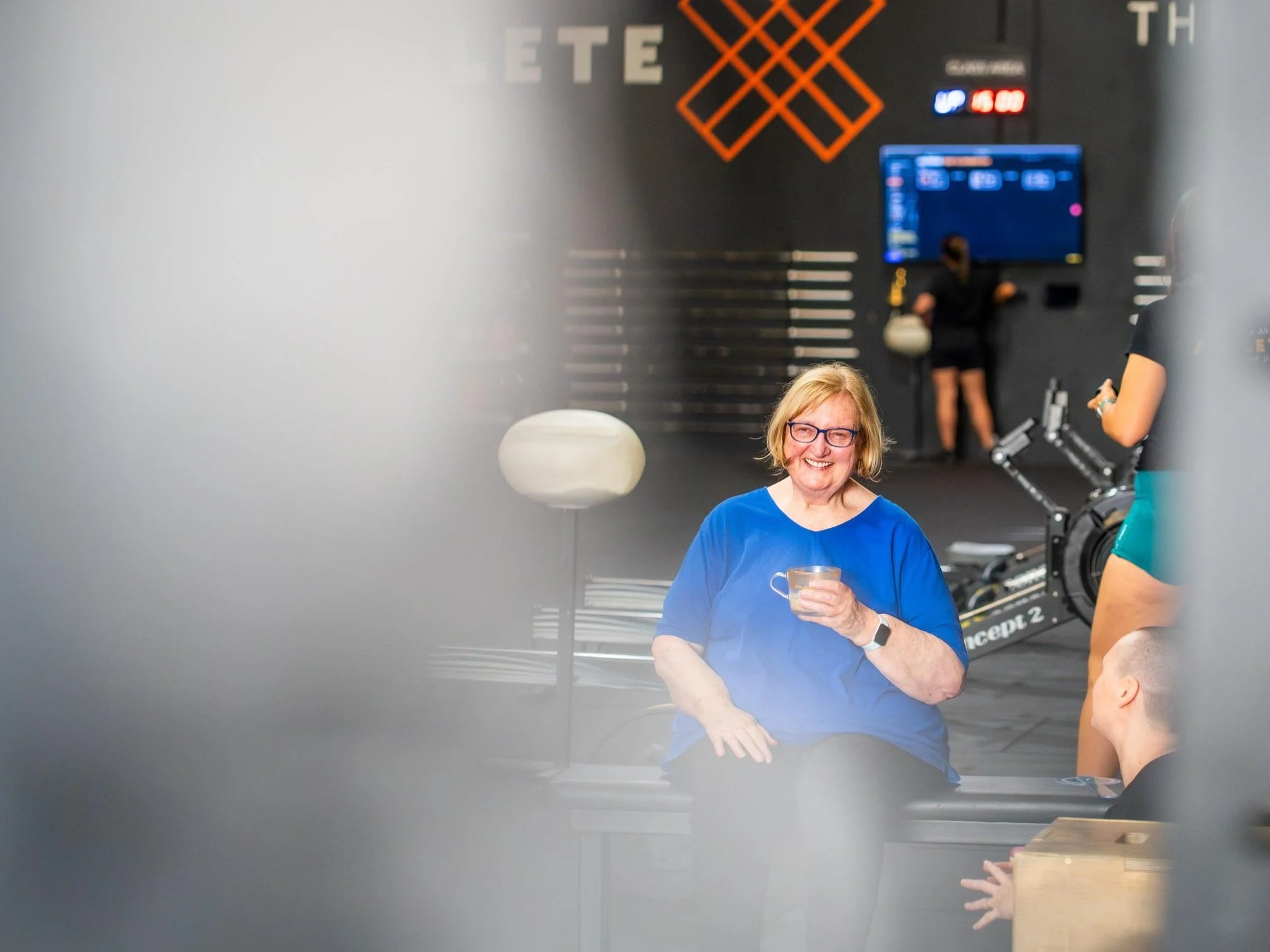 Clare sits on a bench wearing a blue tshirt, holding. a cup of coffee and smiling. In the background is the gym warehouse space with barbells on the wall.