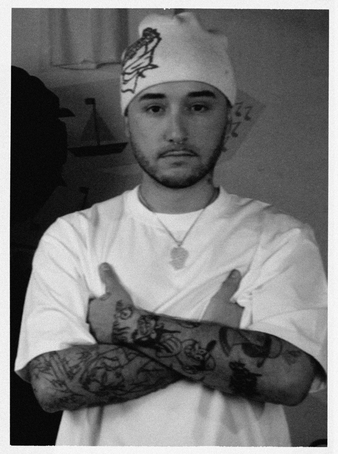 Jeune homme avec un bandana sur la tête, portant un t-shirt blanc et une chaîne en collier, montrant ses bras tatoués croisés devant lui. Fond simple.