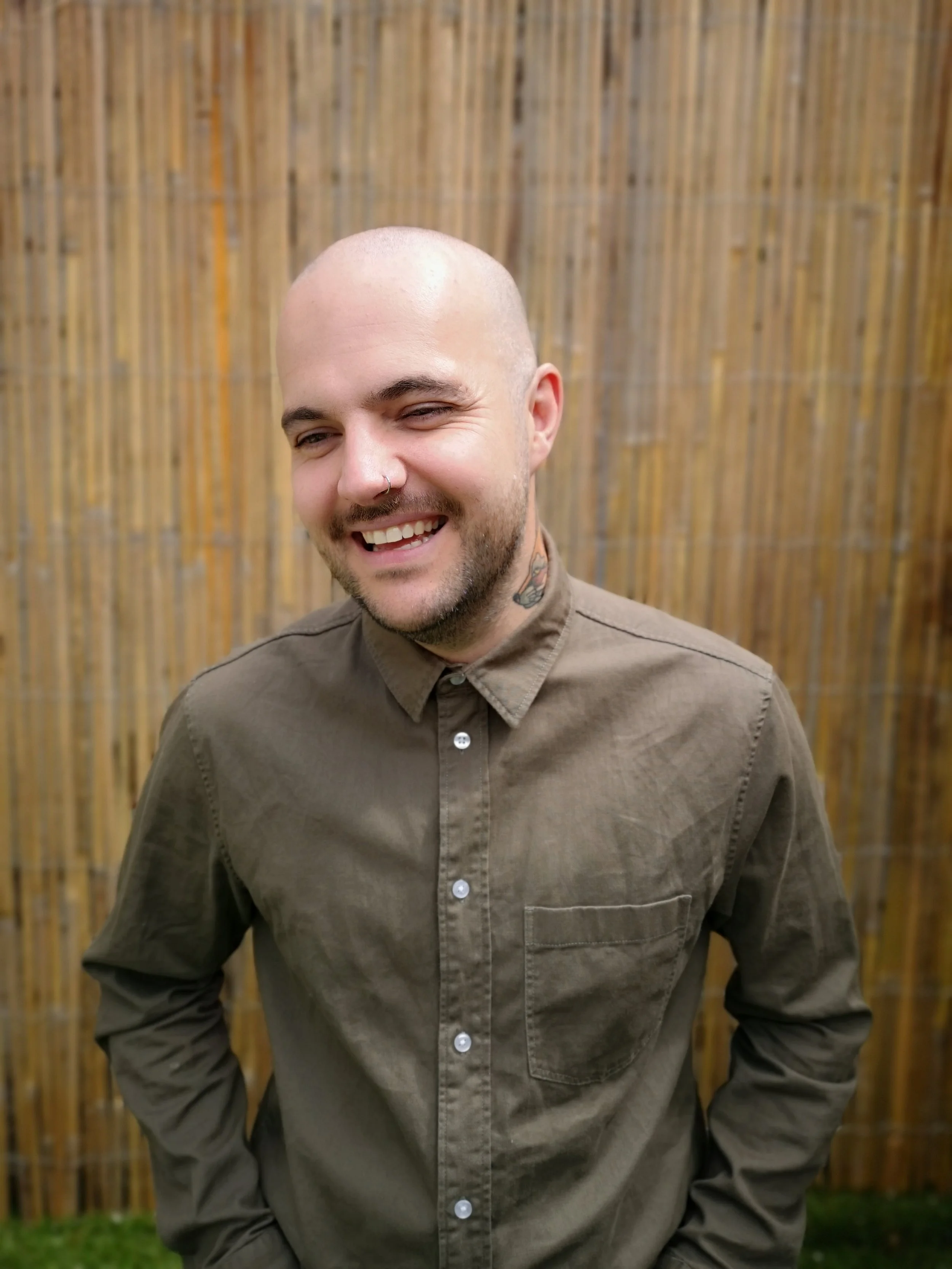 A smiling, bald man with a nose piercing and tattoos, wearing a brown button-up shirt, standing in front of a bamboo fence.