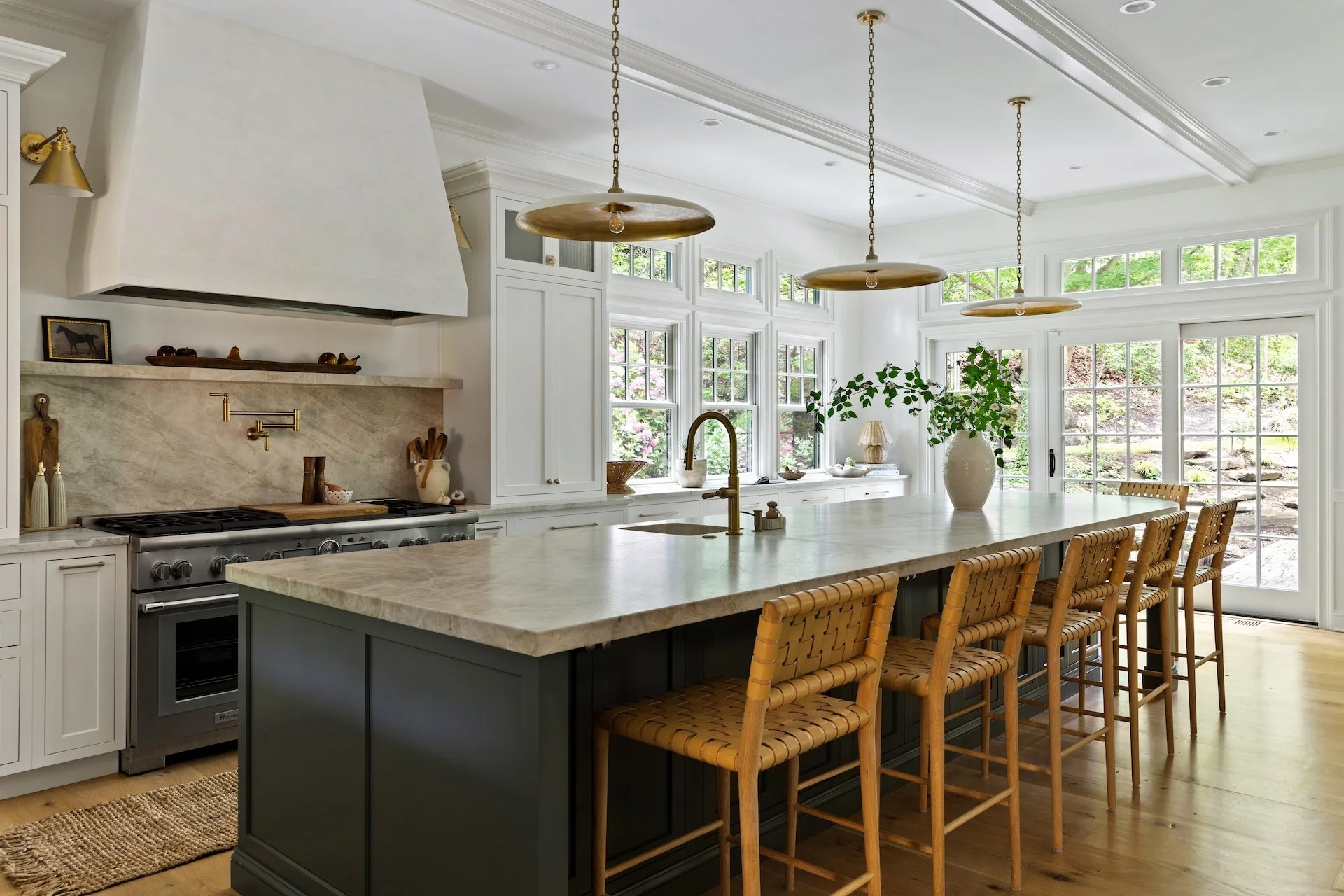 Custom kitchen island with marble countertop, brass pendant lights, woven bar stools, and large windows overlooking a landscaped yard