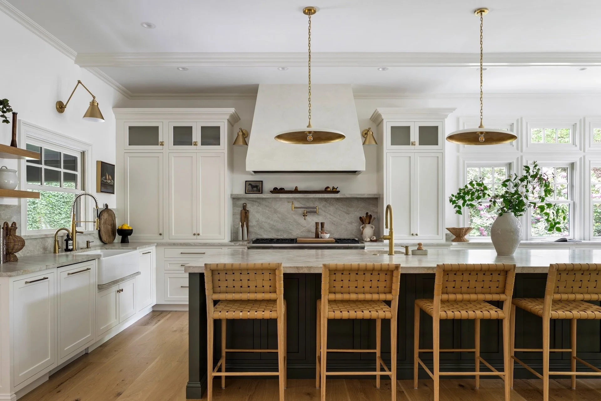 Bright custom kitchen with white cabinetry, marble countertops, brass lighting, and a dark island with woven counter stools