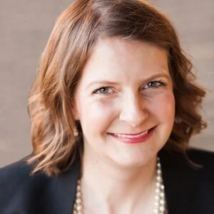 Close-up of a smiling woman with shoulder-length brown hair, wearing a black blazer and a pearl necklace, against a neutral background.