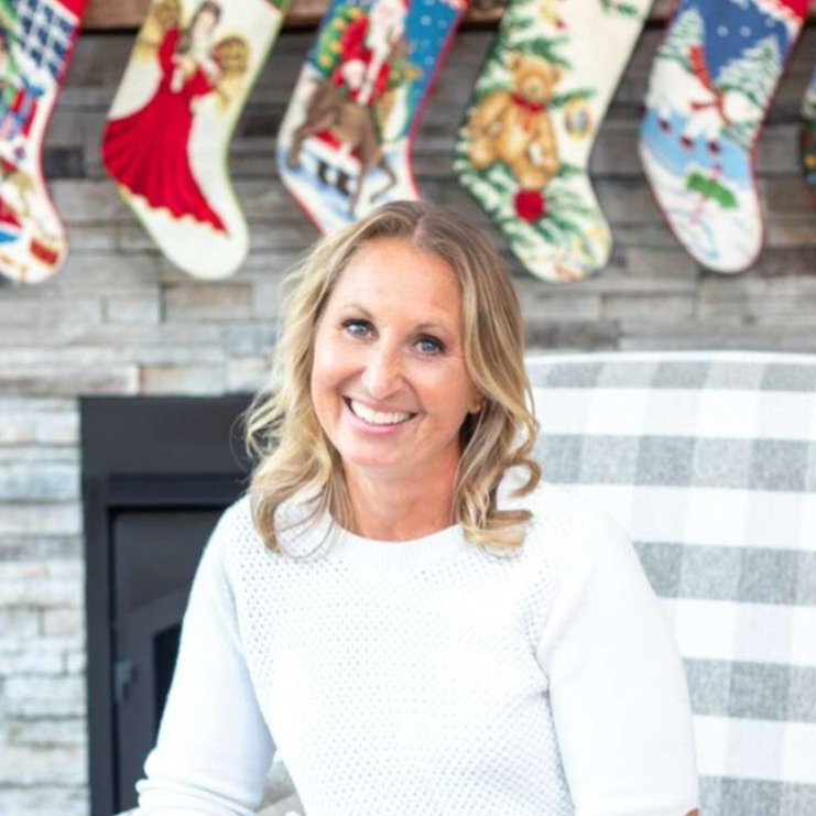 A smiling woman with blonde hair in a white sweater sitting in front of a fireplace decorated with Christmas stockings and ornaments.