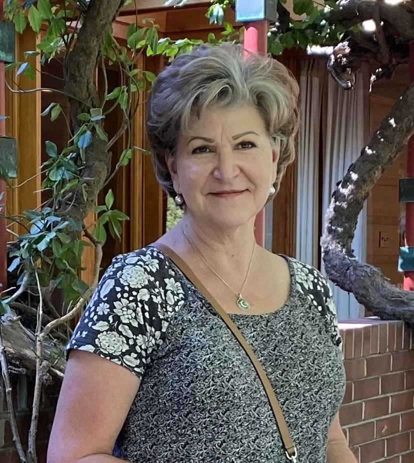 A woman with short gray hair standing outside near trees, wearing a floral-patterned blouse and jewelry.