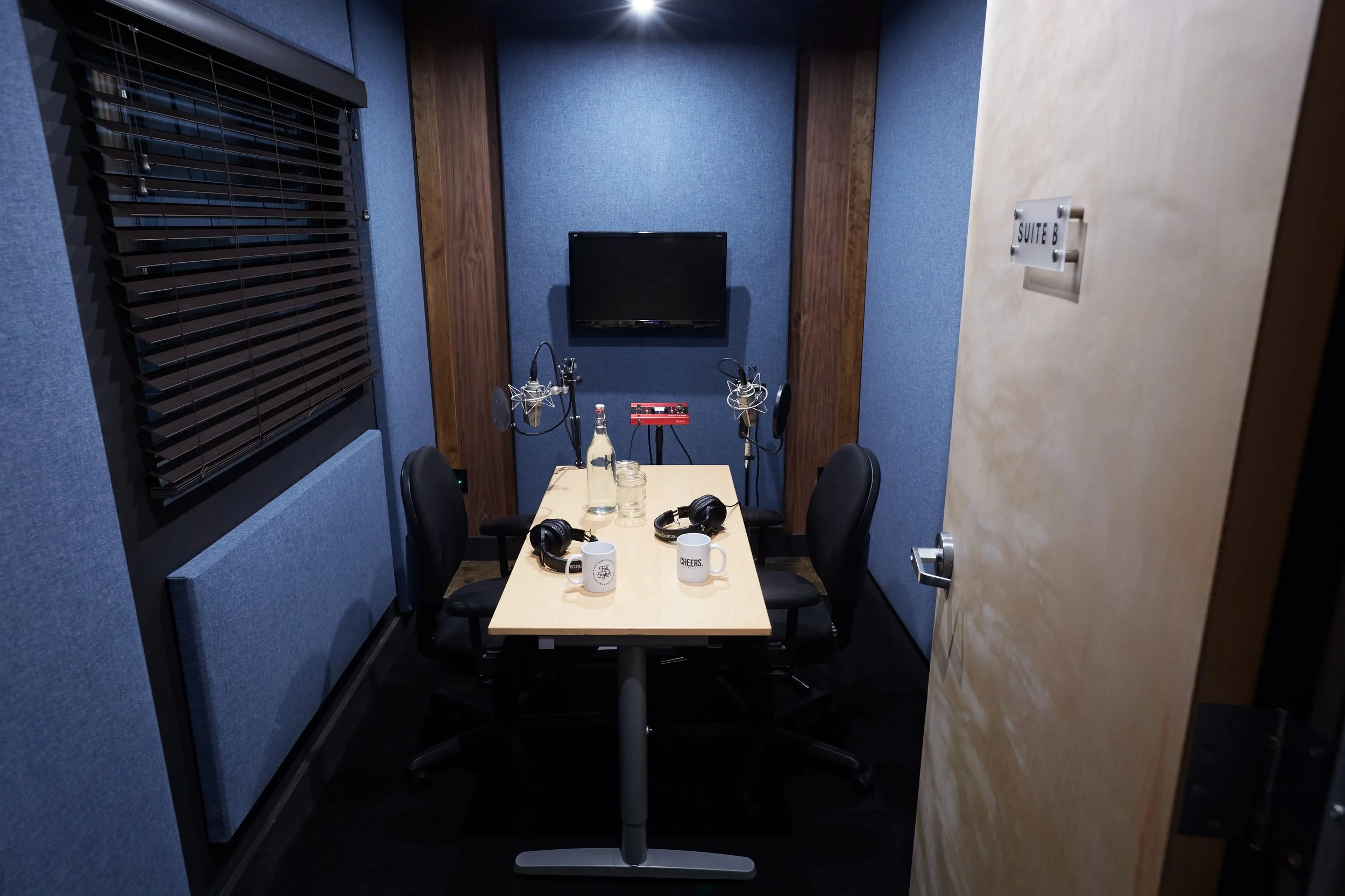 Recording studio with a table, four chairs, microphones, headphones, mugs, glass bottles, and a wall-mounted TV in a blue and wood-paneled room.