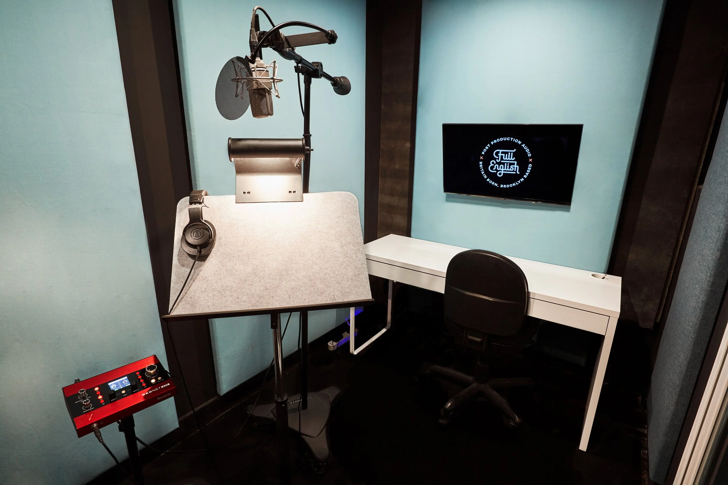 Recording studio booth with microphone, pop filter, headphones, and a small sound mixer, adjacent to a white desk with a black office chair and a wall-mounted TV screen.