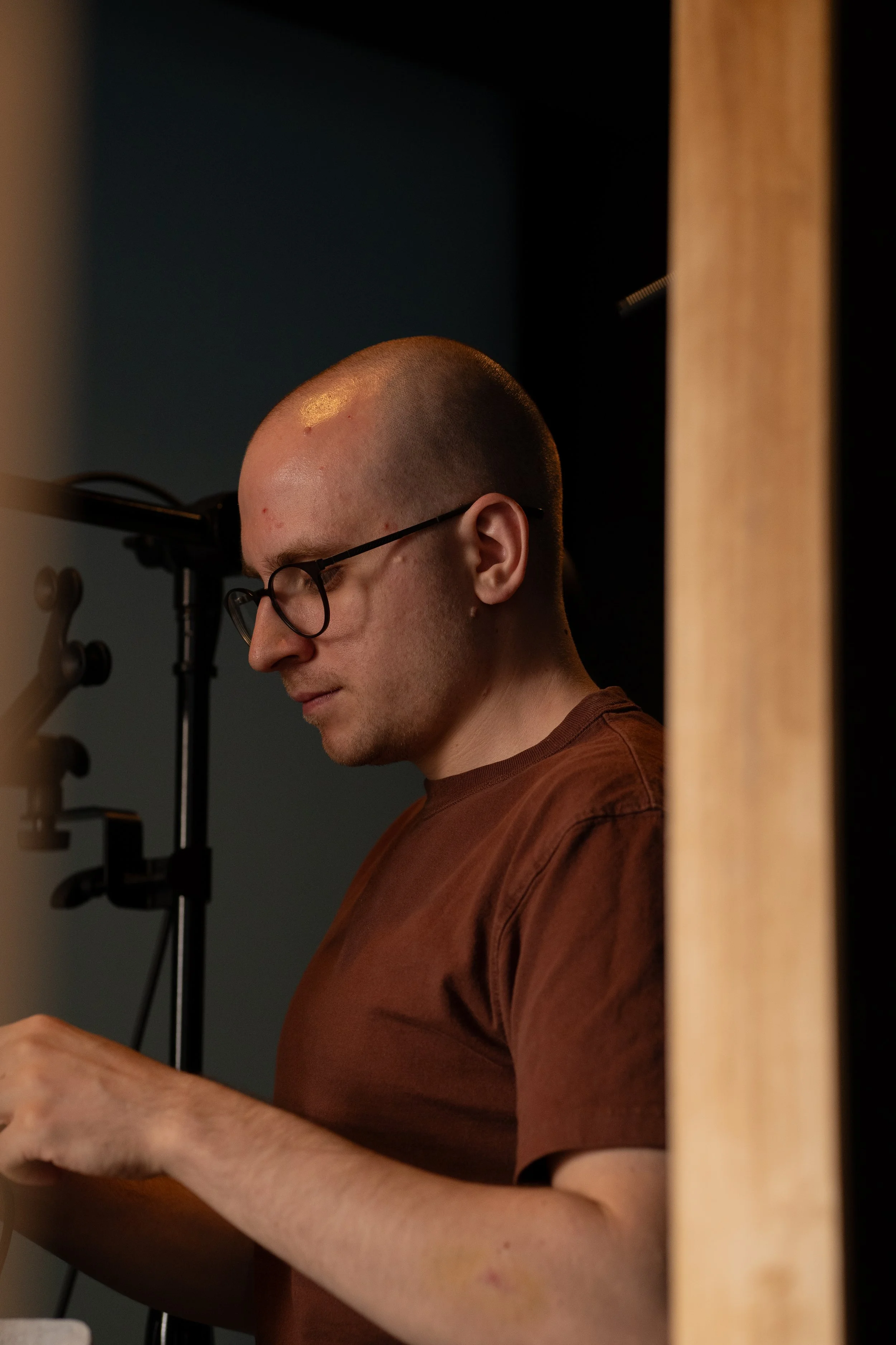 A man with glasses and a shaved head, wearing a brown t-shirt, looking down or reading while standing near a wooden piece of furniture, with a dark background.