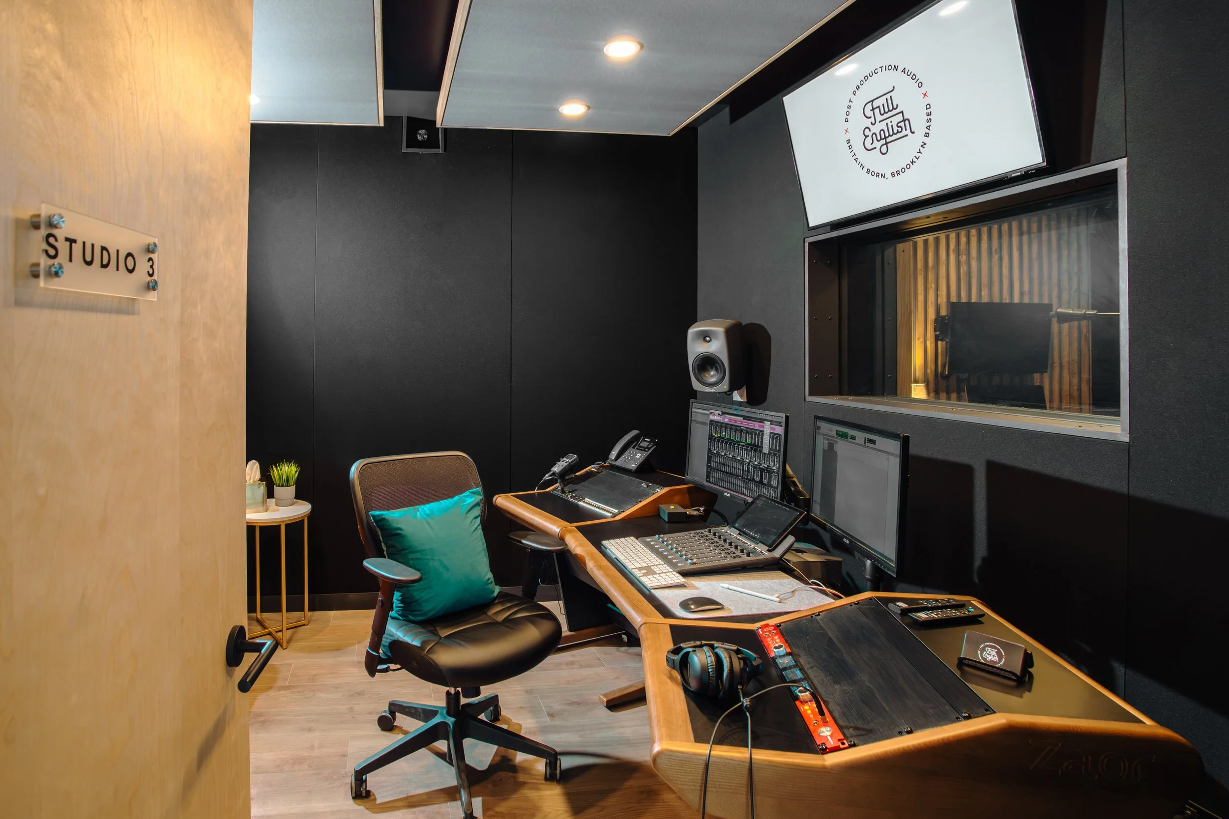Recording studio with black soundproof walls, wooden control desk with audio equipment, multiple computer monitors, a chair with teal cushion, and a window to an adjacent recording booth.
