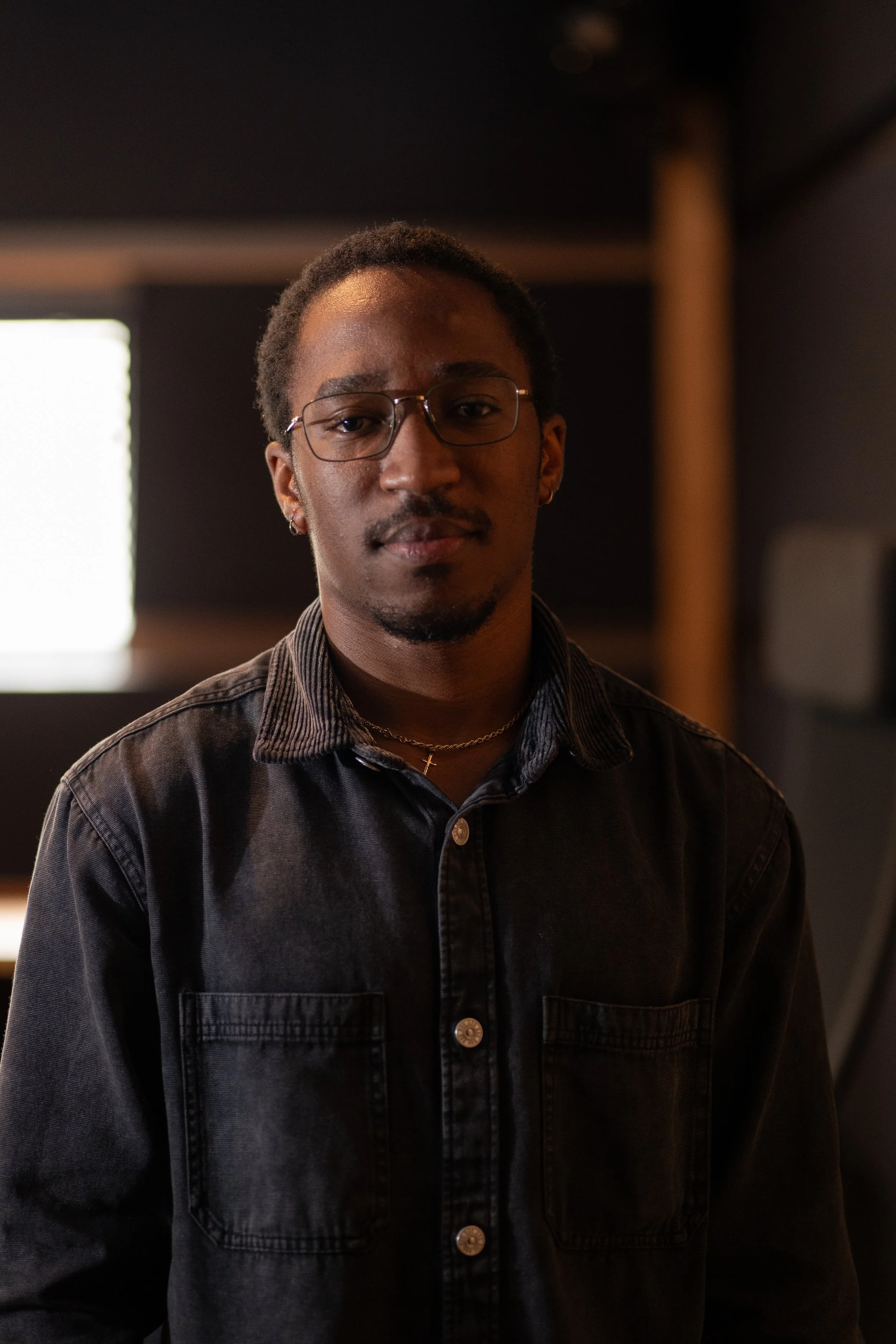 Portrait of a young man wearing glasses, a black denim shirt, and a gold cross necklace, standing indoors with a dark background and light coming from a window.