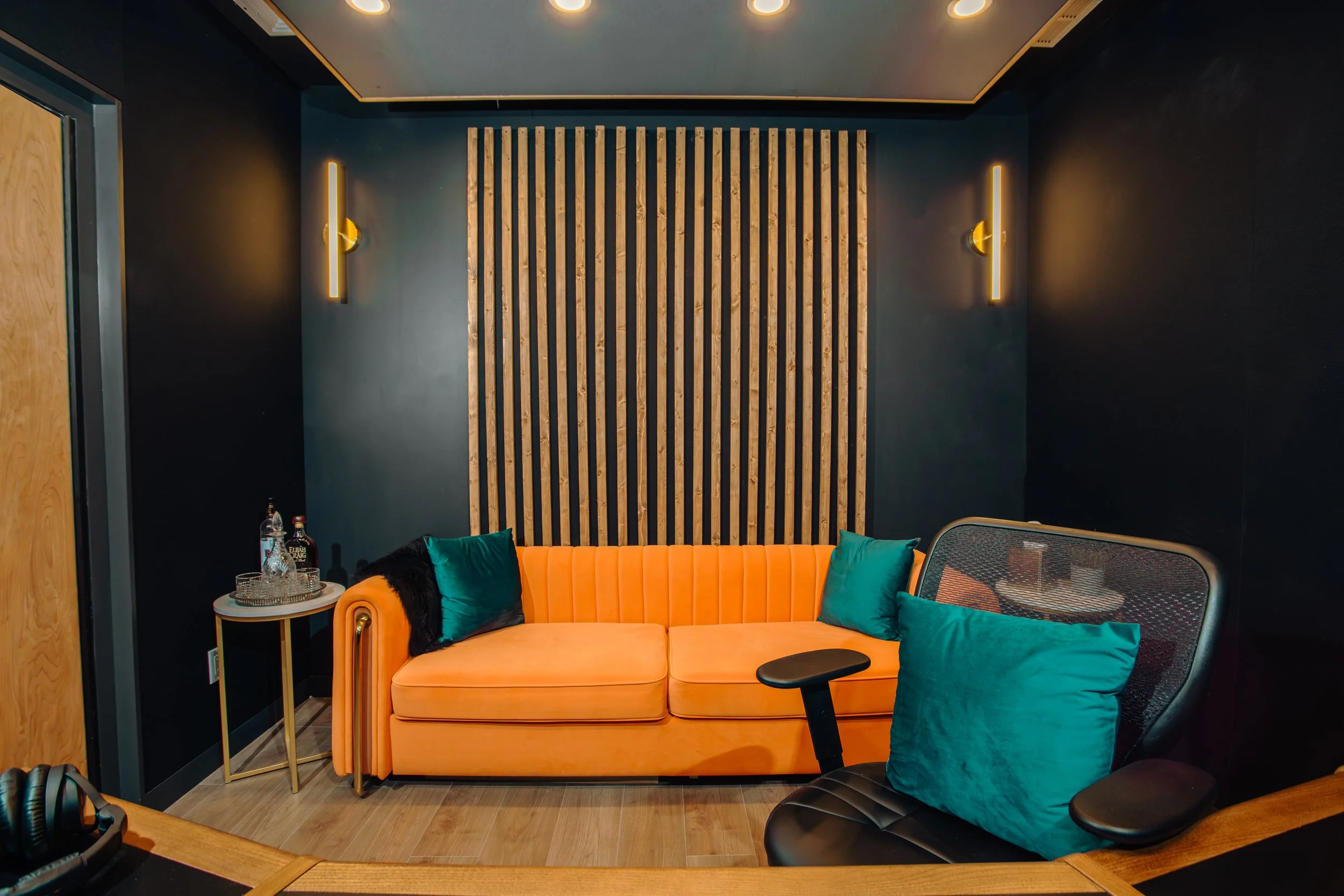 A modern recording or radio studio room with a peach-colored sofa, teal pillows, and a black swivel chair. There is a small side table with drinks on it, and a wooden slat wall panel behind the sofa. The room has dark walls, wood flooring, and minima