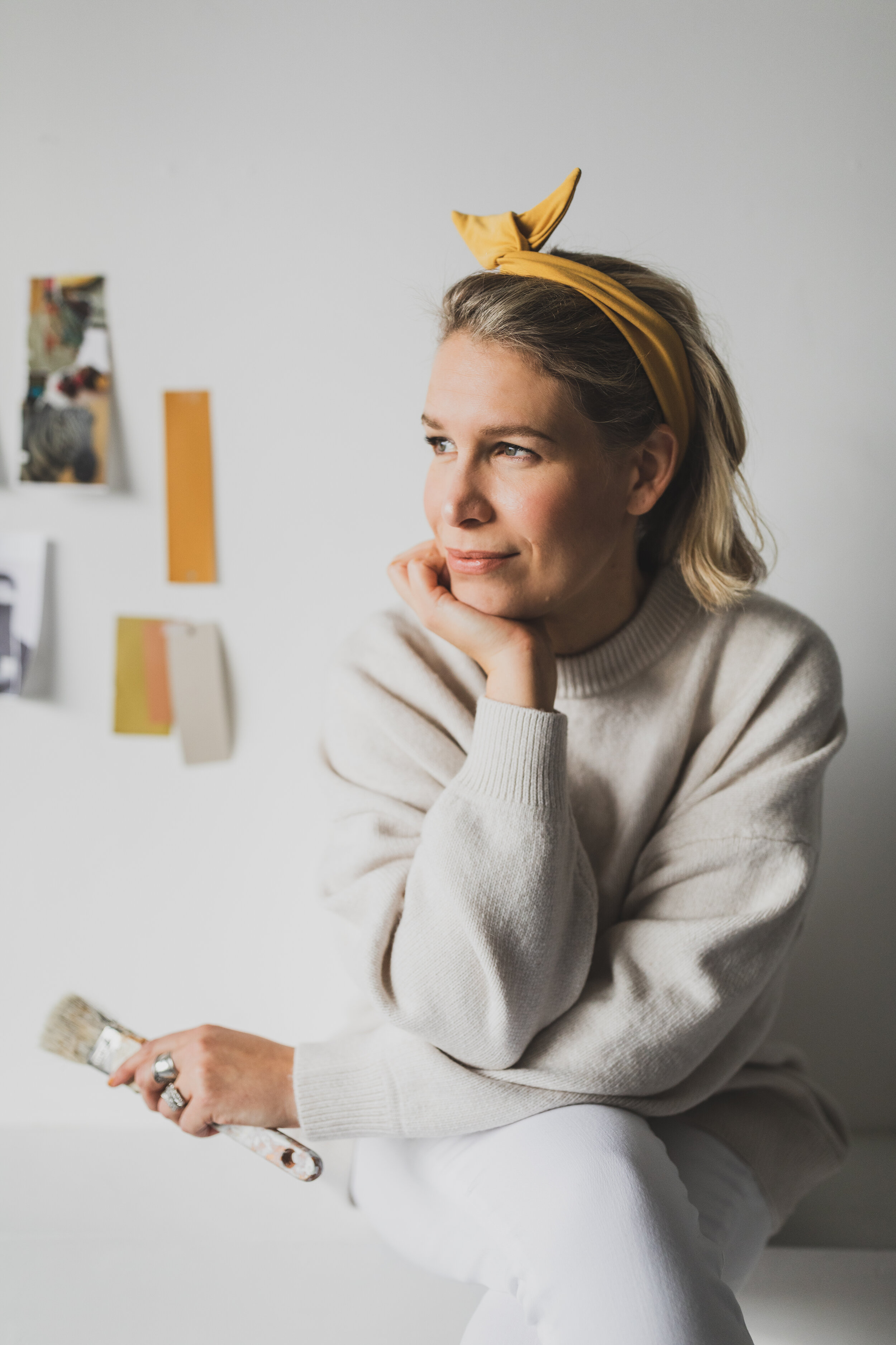 A woman with blonde hair wearing a yellow headband, sitting with her chin resting on her hand, holding a paintbrush in her other hand, in front of a plain white wall with colorful photos or papers pinned to it.