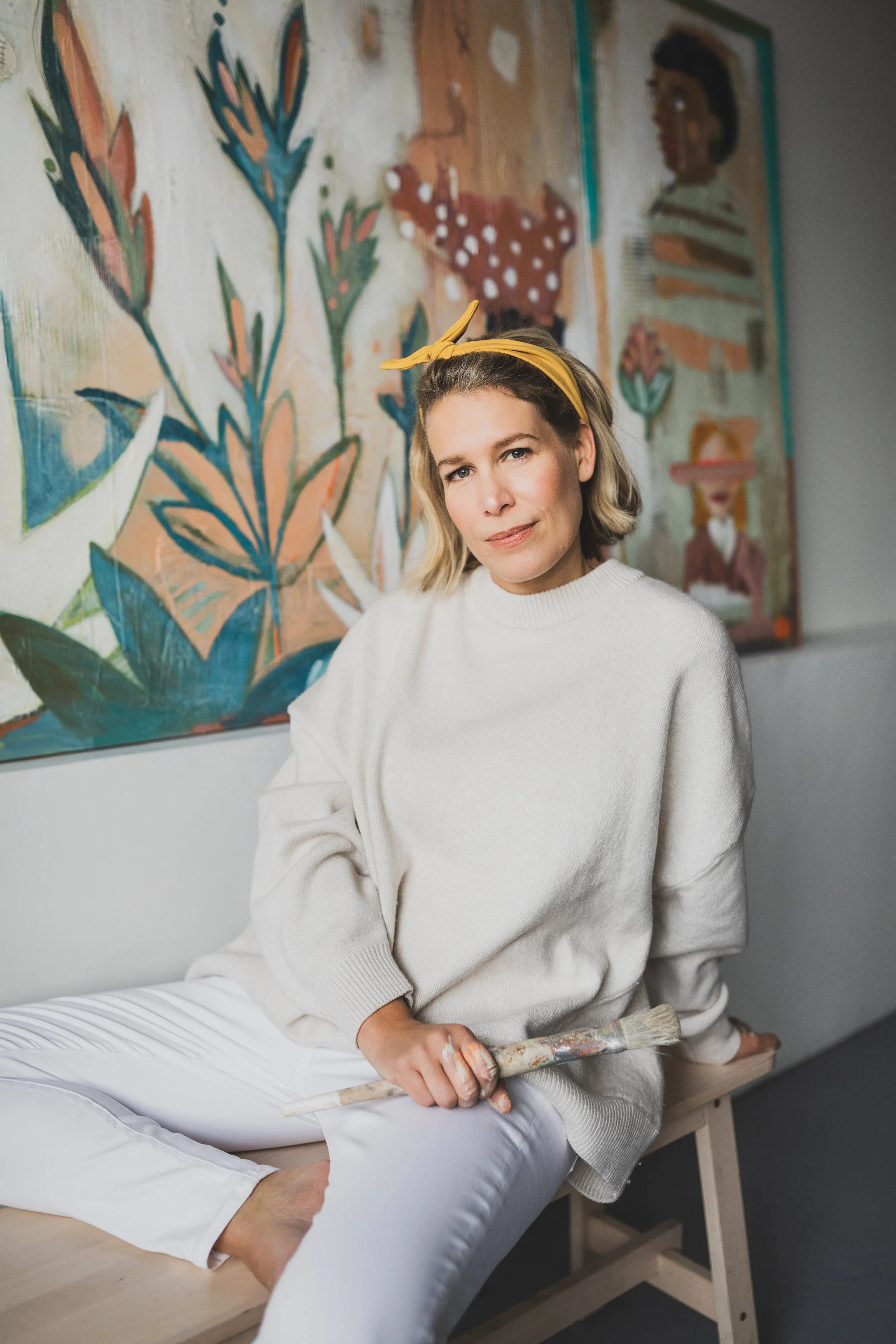 A woman sitting on a wooden bench holding a paintbrush, wearing a cream sweater and white pants, with a yellow headband, in front of a colorful painting of stylized figures and plants.