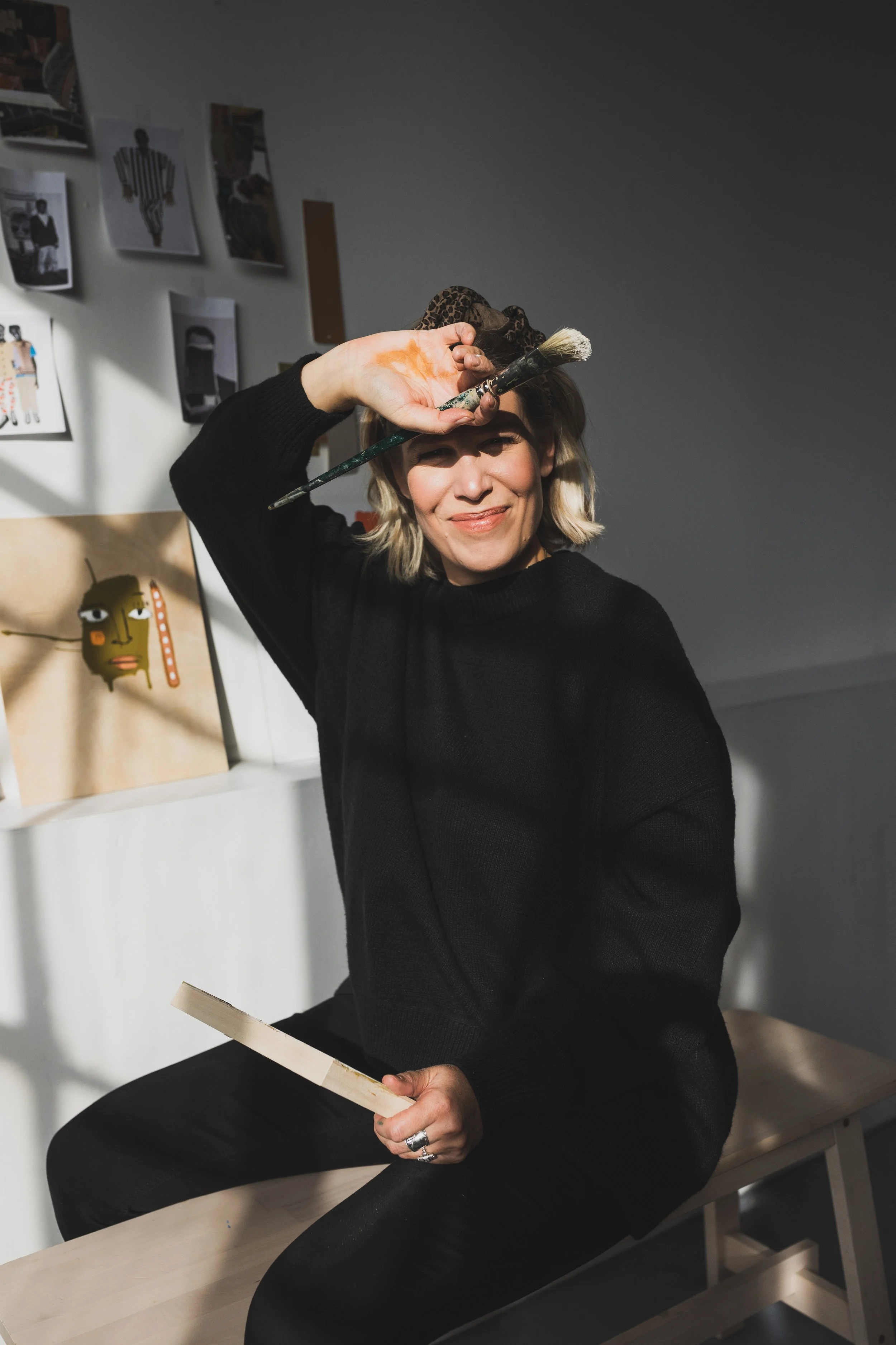 A woman seated on a bench, smiling, holding a paintbrush in one hand and a wooden object in the other. She is wearing a black sweater with sunlight casting shadows on her face. Behind her, there are artworks pinned on the white wall, including a colorful face painting and pictures.