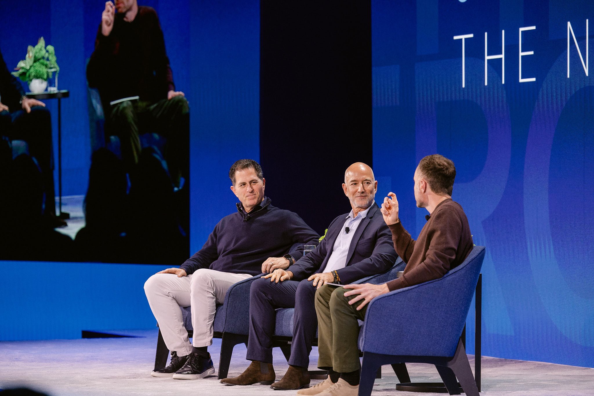 Three men seated on blue chairs on stage during a panel discussion, with a blue background and a large screen behind them displaying a mirror image of the scene.
