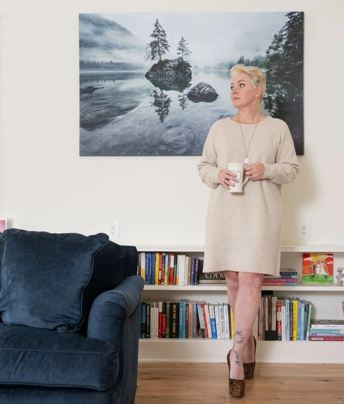 A woman with short blonde hair and earrings, wearing a beige cable-knit sweater, standing by a window with cream curtains, looking at the camera.