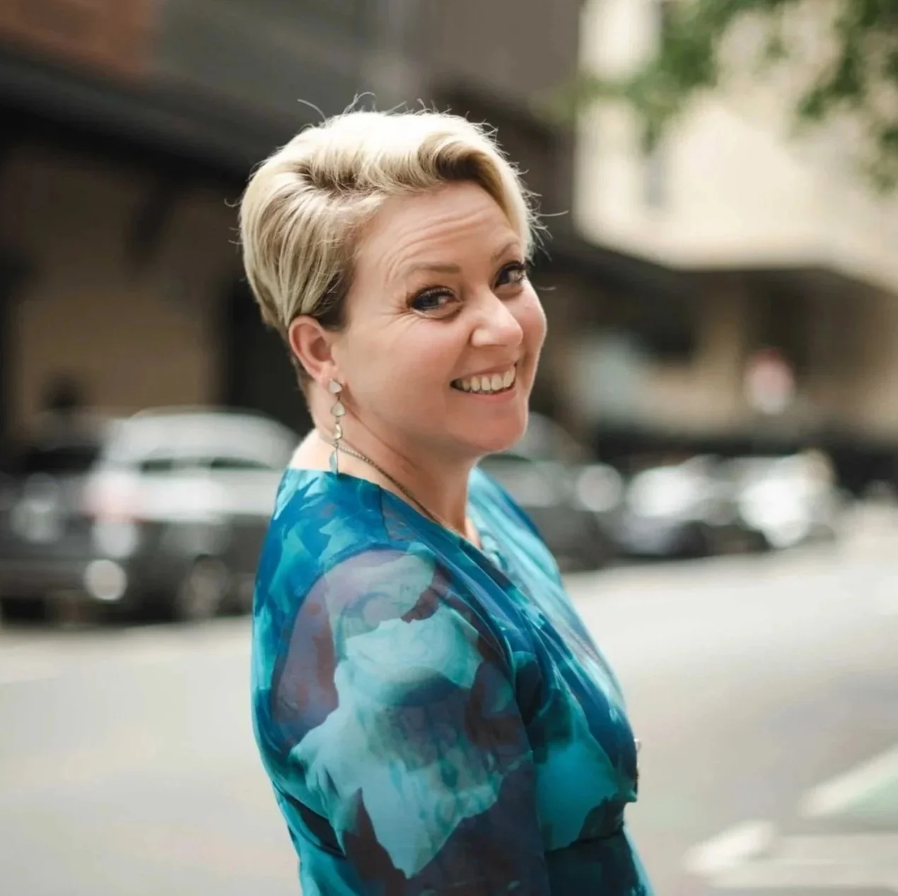 Smiling woman with short blonde hair wearing a blue patterned dress standing on city street with cars and trees in background.