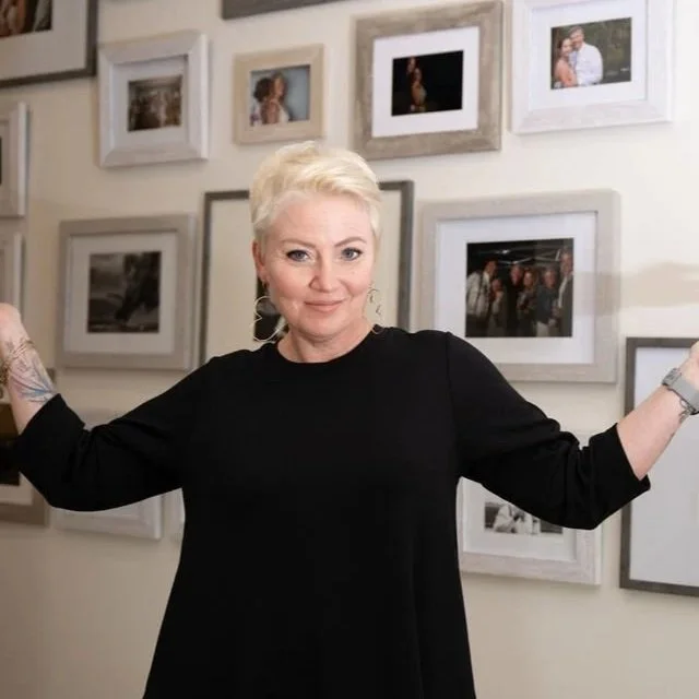 A woman with short blonde hair wearing a black top, standing in front of a wall decorated with framed photographs, with her arms extended outward.