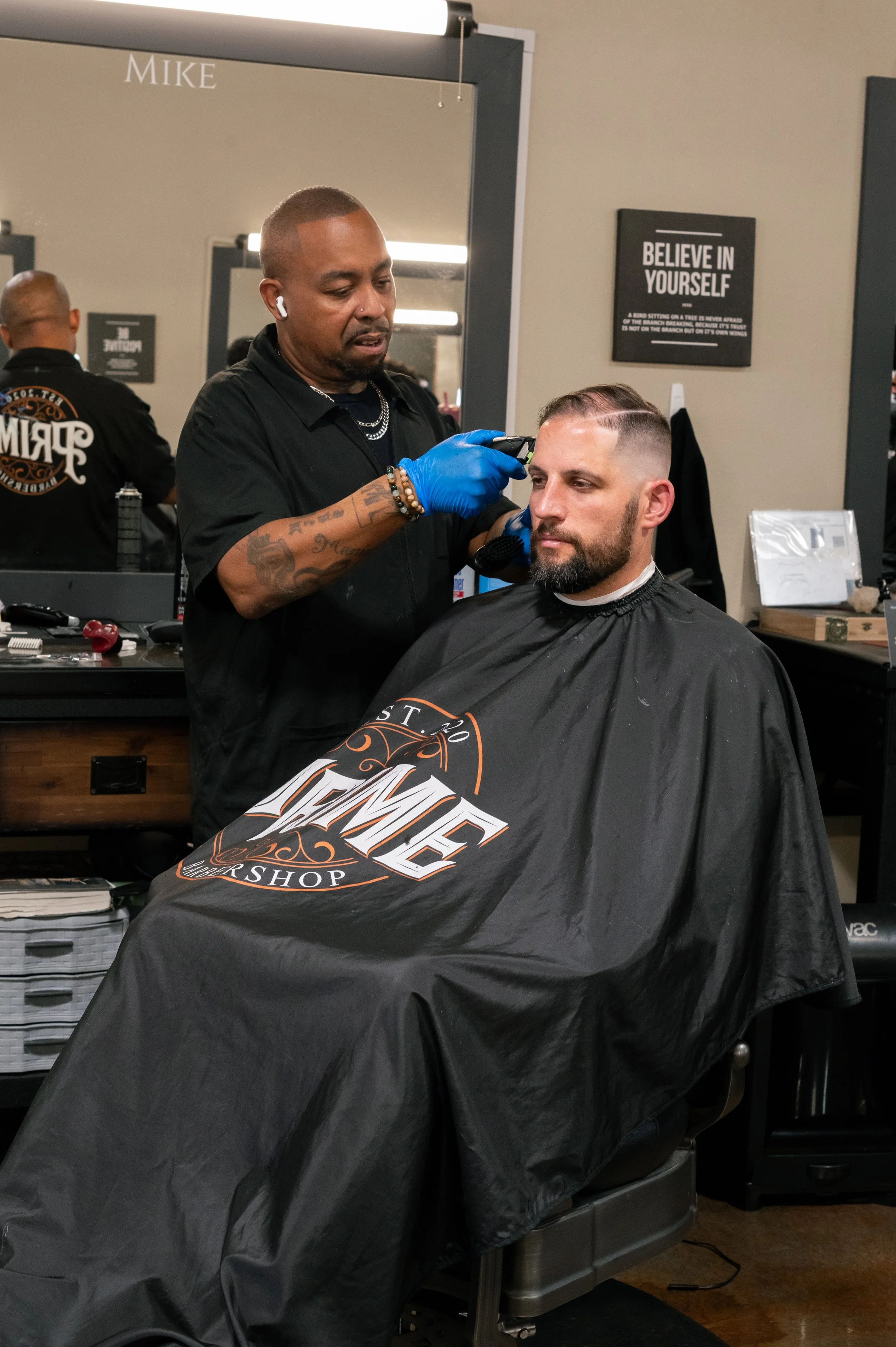 A barber gives a man a haircut in a barber shop, with a large mirror behind them. The barber is using clippers on the man's beard, who is sitting under a black barber cape with the shop's logo.
