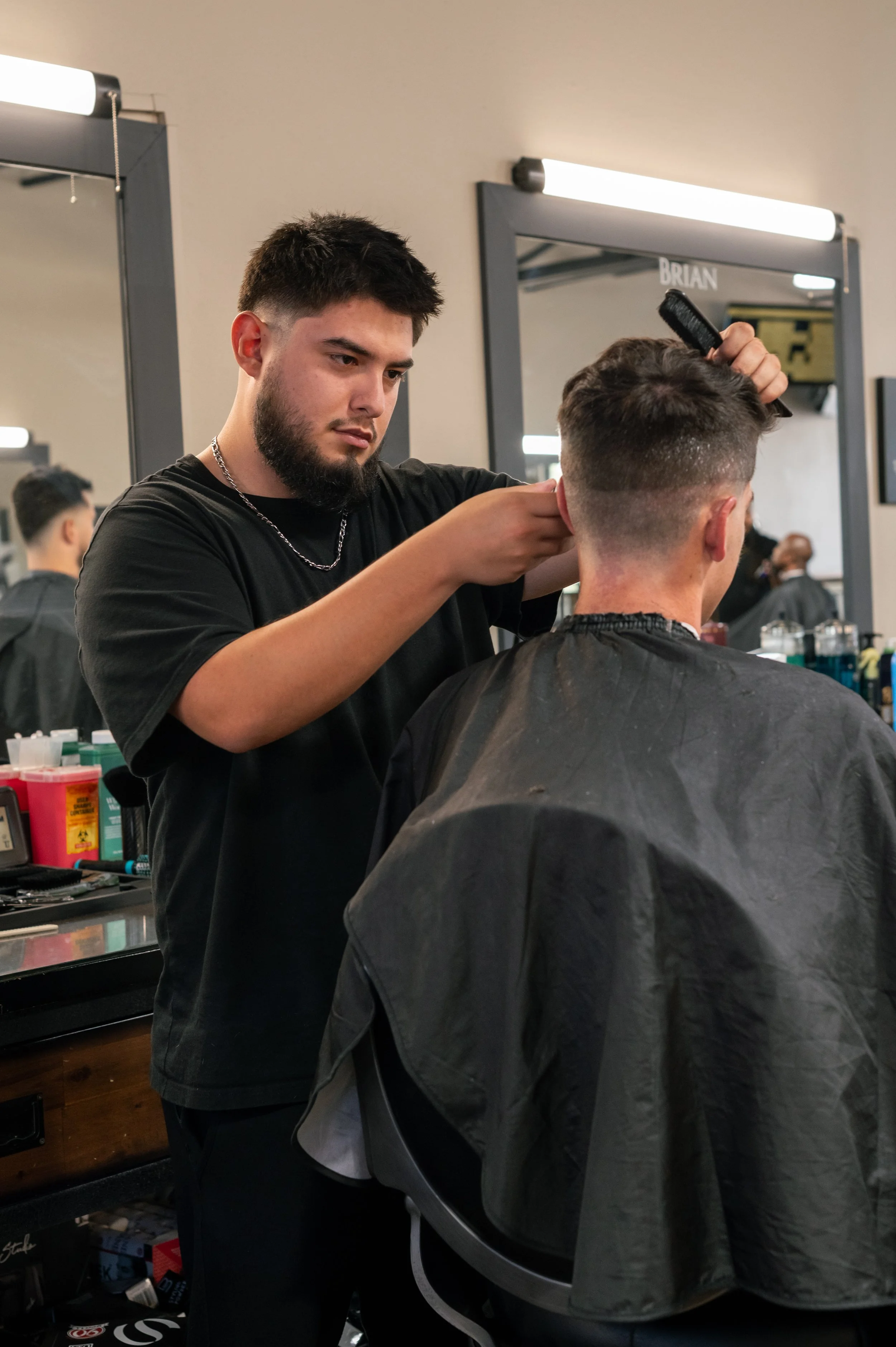 A barber with dark hair and beard cutting a male customer's hair in a barber shop, with mirrors and hair care products in the background.