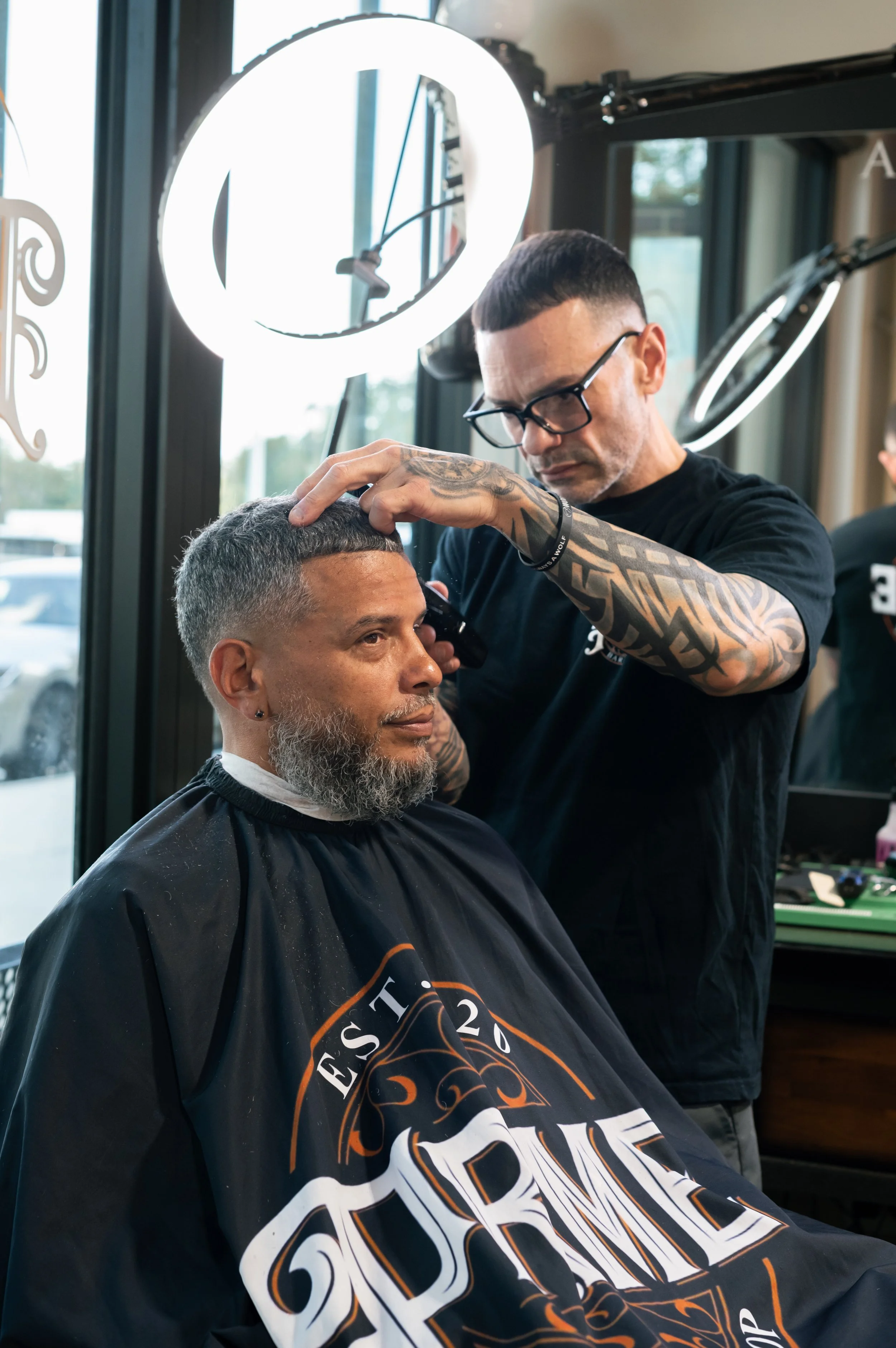 A man getting a haircut at a barbershop, wearing glasses and a black cape with orange and white design, while a barber with tattoos and glasses cuts his hair.