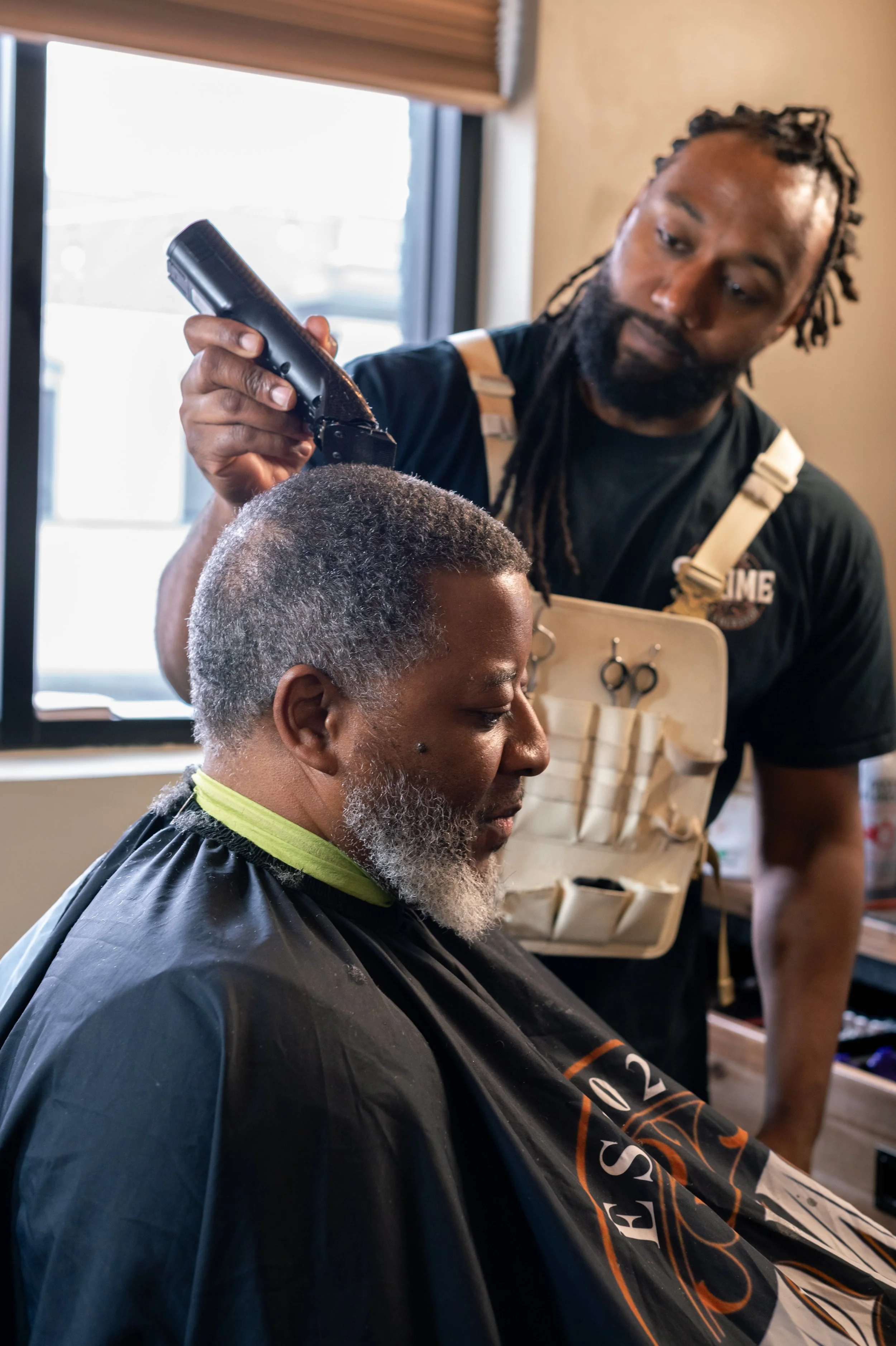 A man with gray hair and beard, wearing a black barber cape, sitting in a barber chair, getting a haircut from a bearded barber, who is holding scissors and a comb near the man's head inside a barbershop.