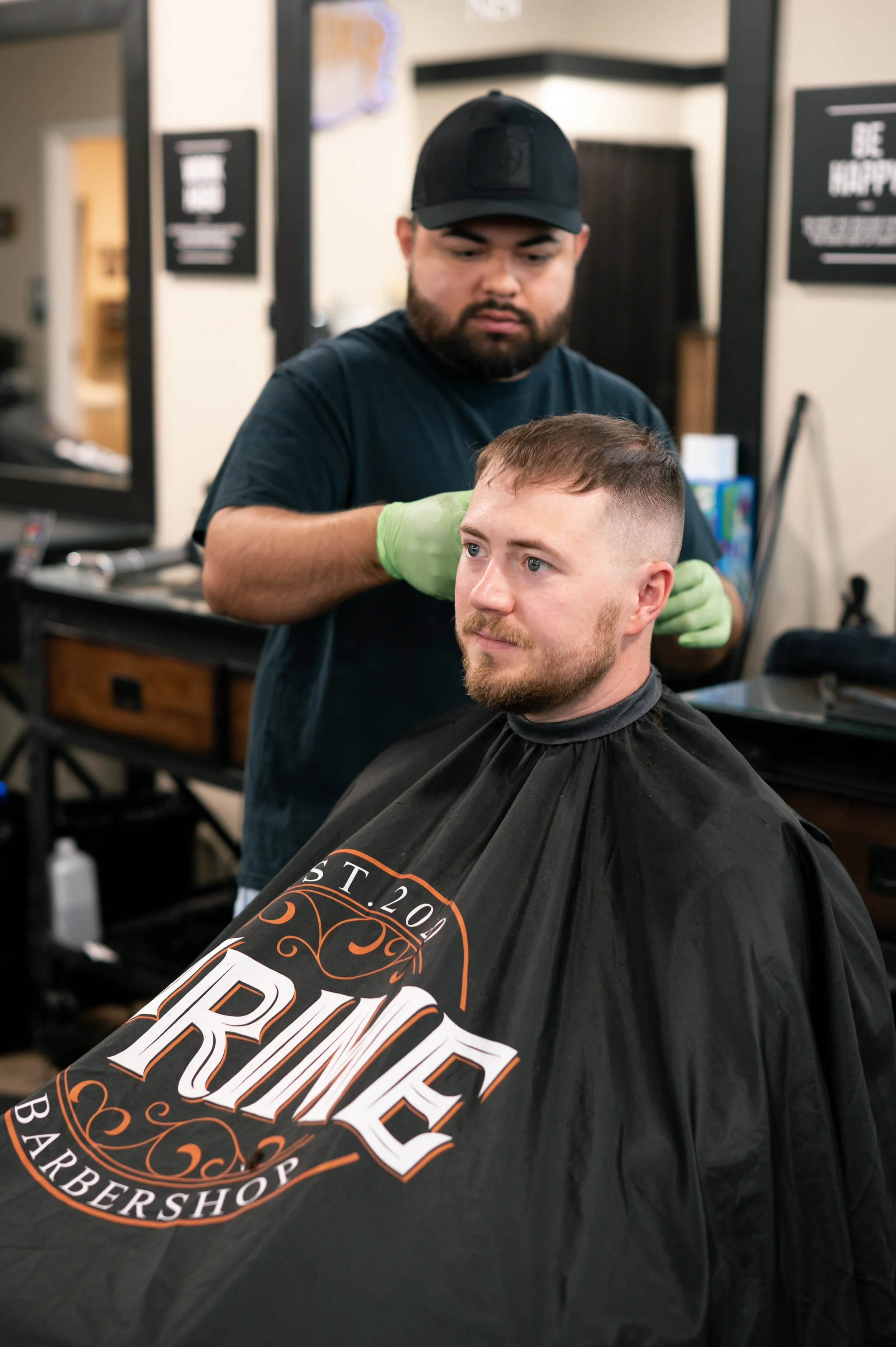 A man with a beard getting a haircut in a barbershop, wearing a black barber cape with the shop's logo.
