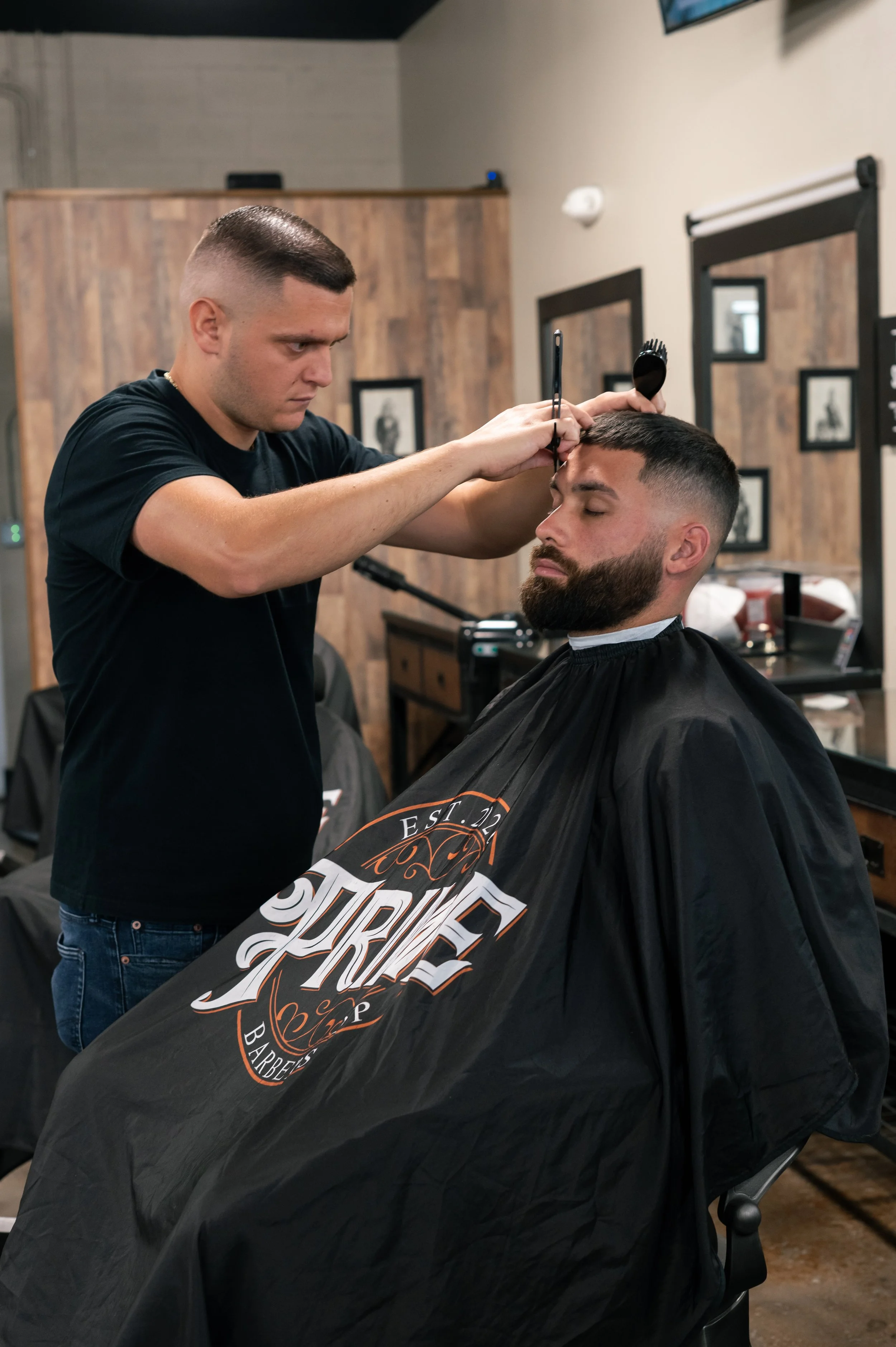 A barber in a black T-shirt cutting and styling a man's hair at a modern barbershop. The client has a beard and a black barber cape with a logo. The background features wooden panels, framed pictures, and salon mirrors.