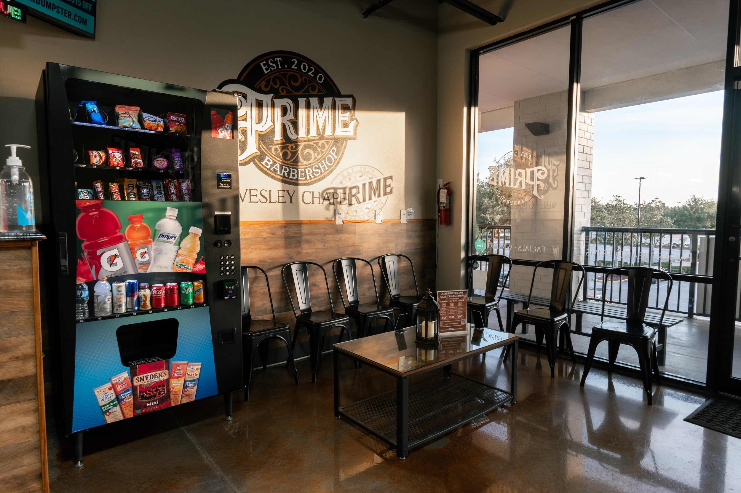 Interior of a barbershop with a vending machine, a wall sign reading 'Prime Barbershop EST. 2020 Wesley Chaperime,' and black chairs around a small table by large windows showing an outdoor parking lot and trees.