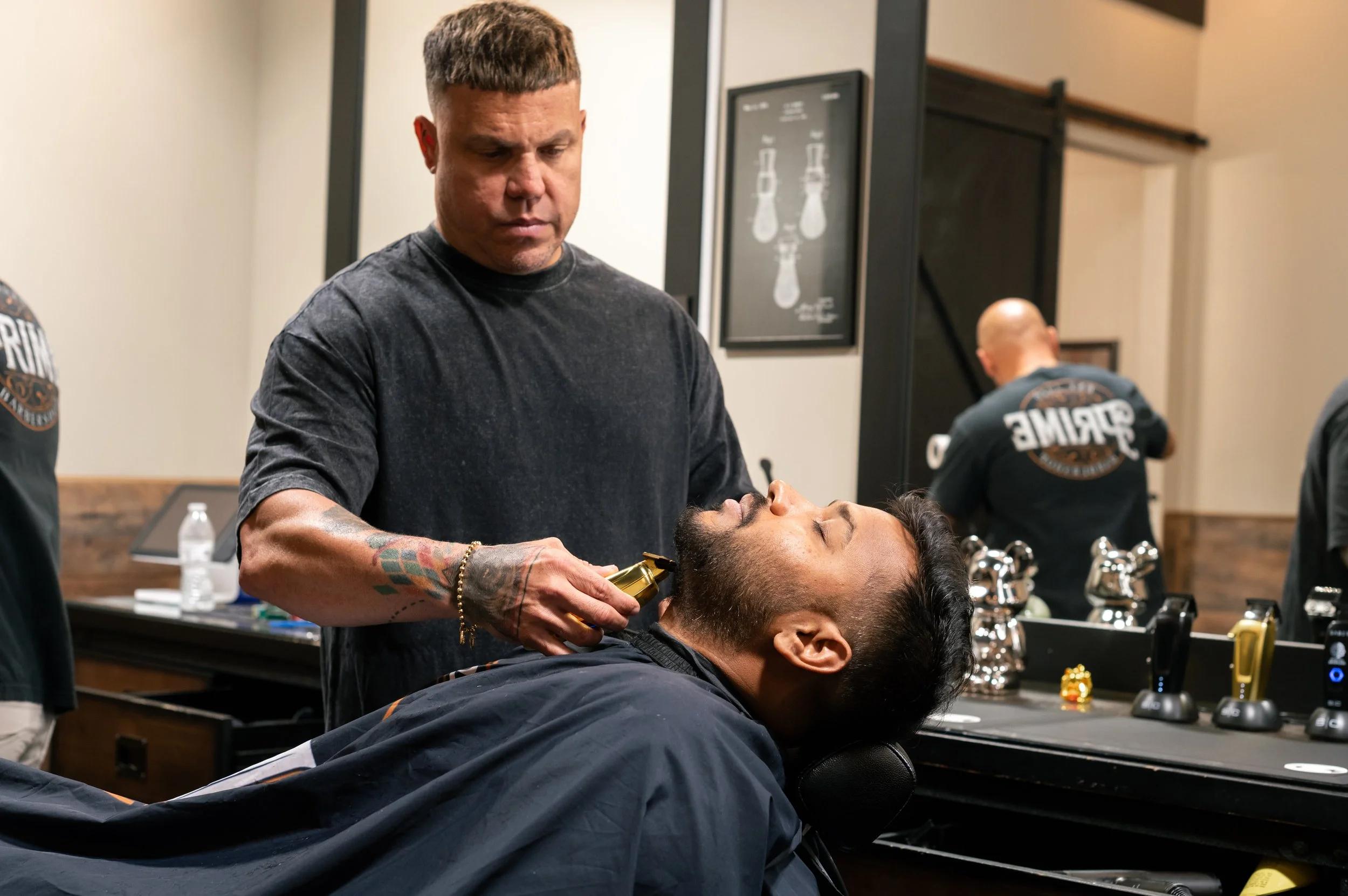 A man getting a shave from a barber in a barbershop. The barber is holding a gold-colored electric shaver, and the man is reclined in a barber chair with a black cape. In the background, there are other barbers working and a framed chart of beard sty