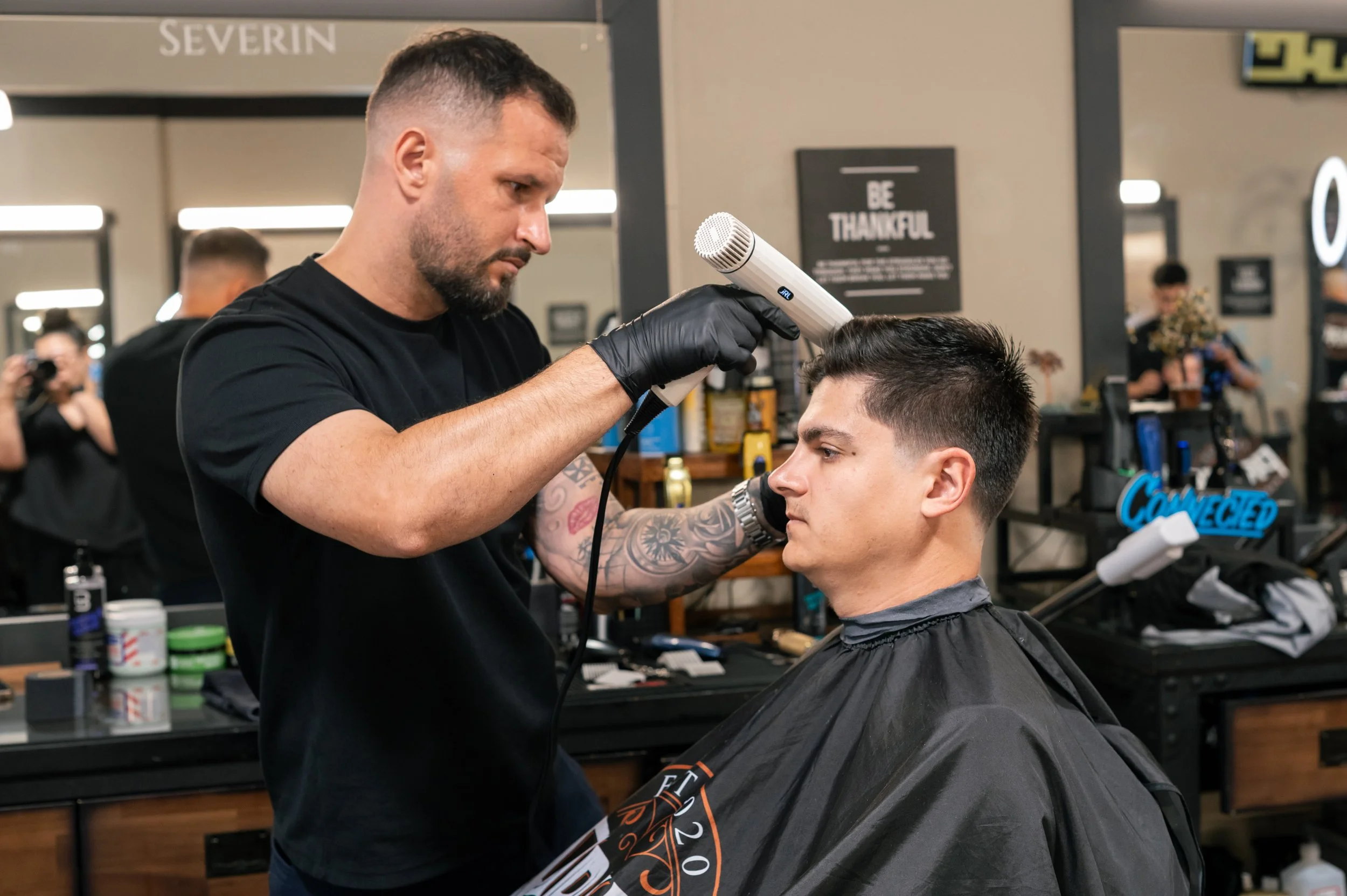 A barber stylishly dries a client's hair with a white handheld blow dryer in a modern barbershop interior.