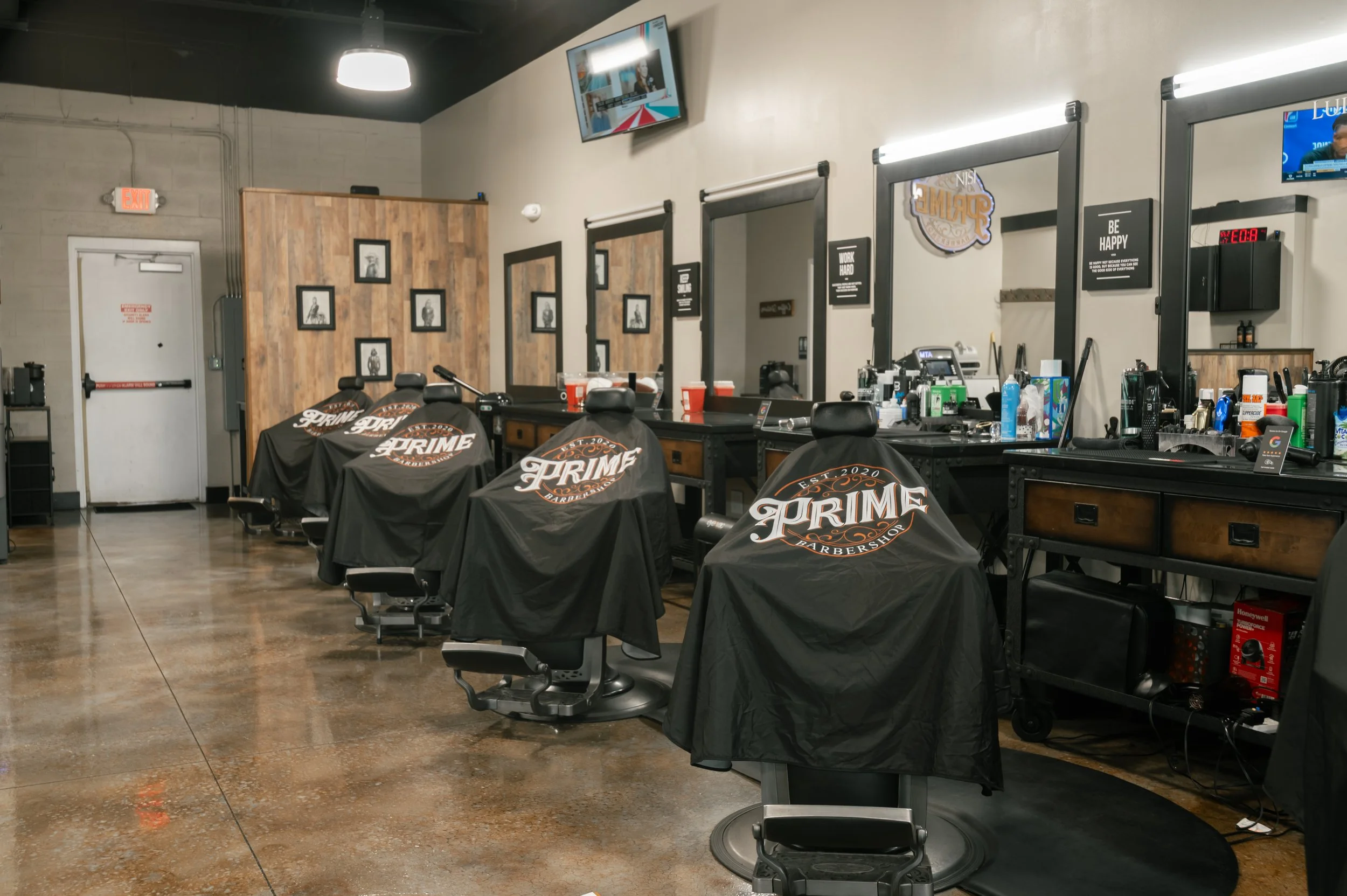 Inside a barbershop with black barber chairs covered in black cloths with the word 'Prime' on them, mirrors on the wall, and hair styling tools and products on the counters. Framed pictures on a wooden wall section, and a TV mounted on the wall.
