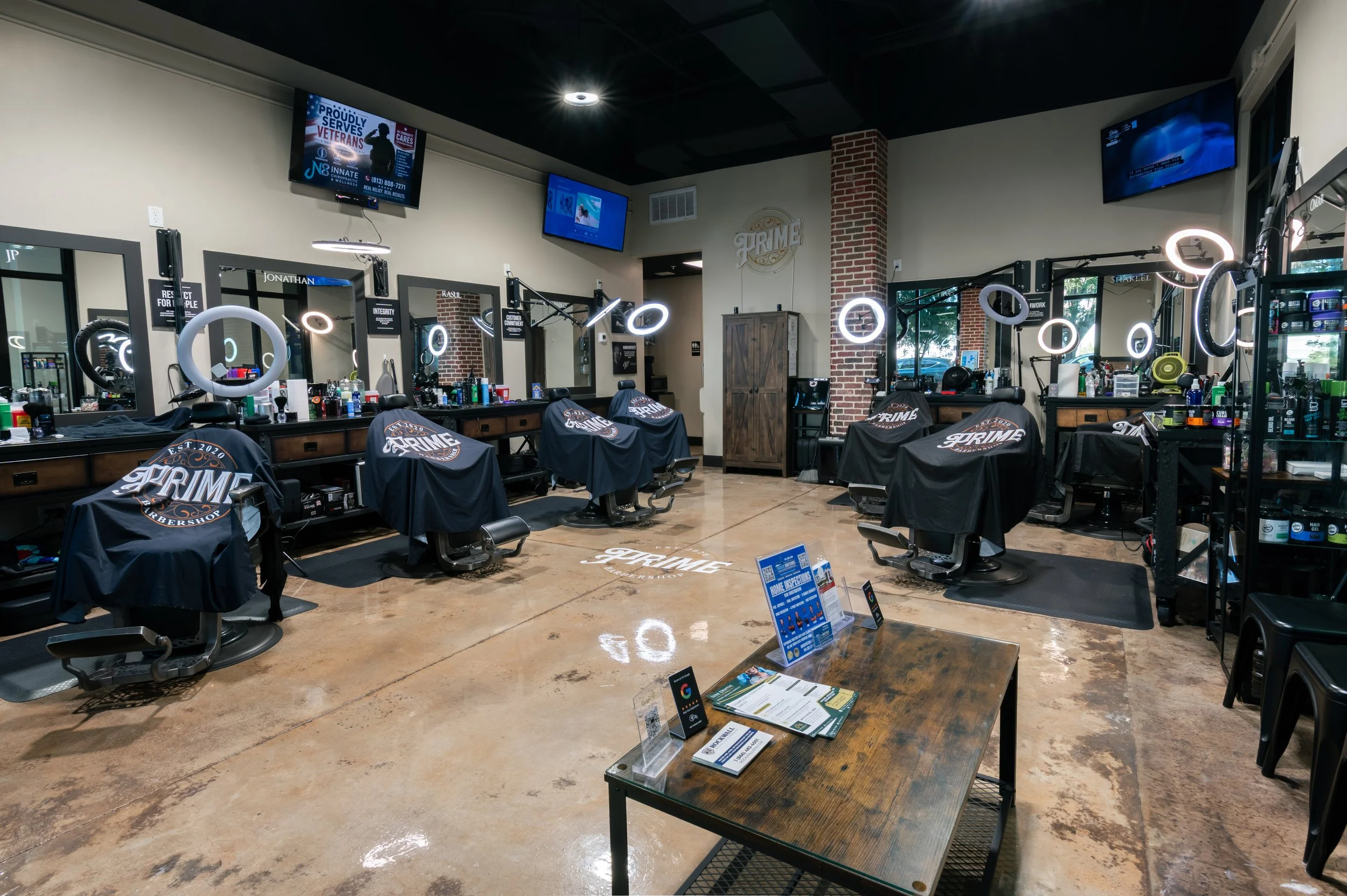 Interior view of a barbershop with multiple styling stations, mirrors, barber chairs covered with black capes, and illuminated rings around some mirrors, with TVs mounted on the wall and products on shelves.