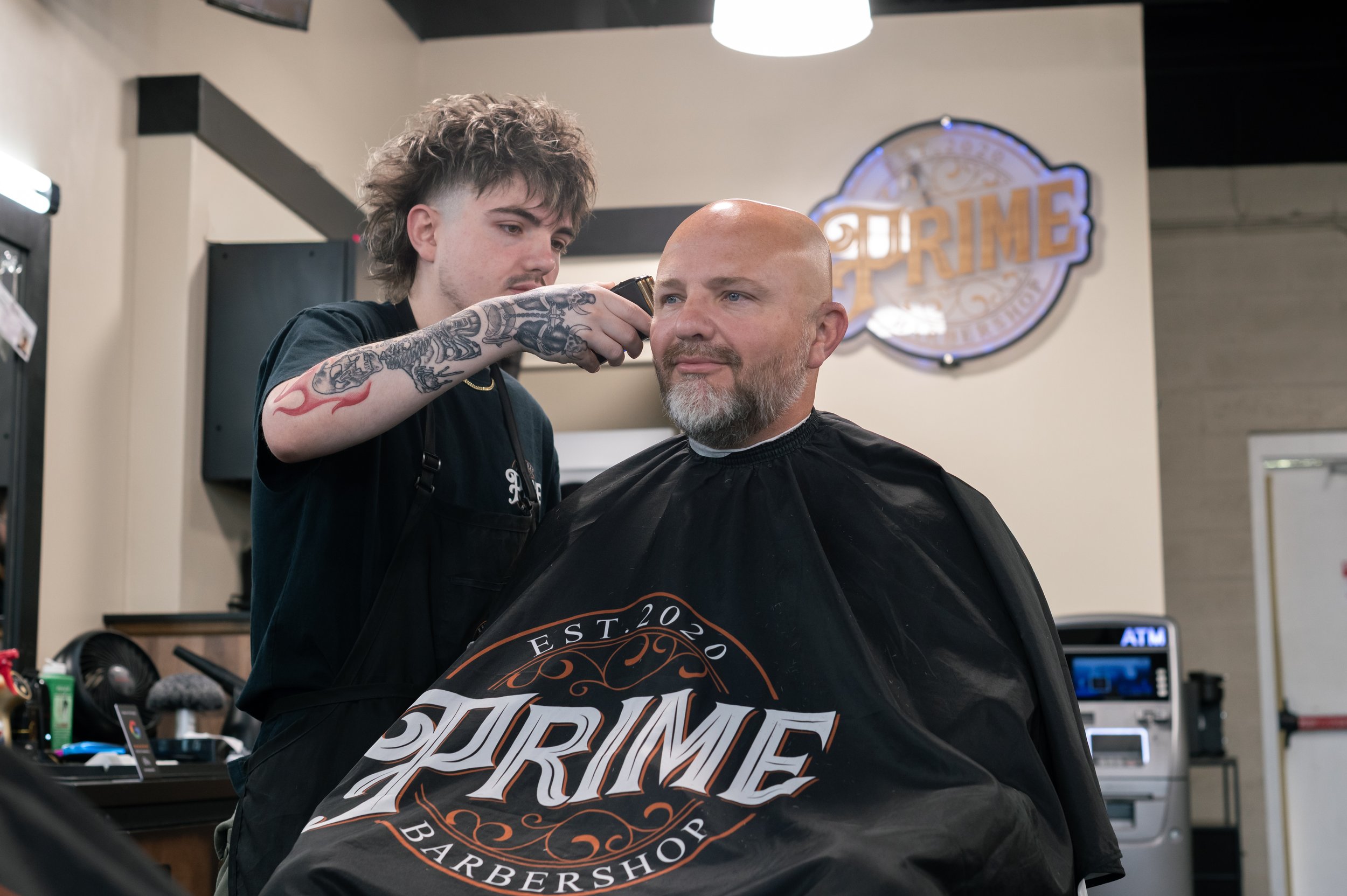 A barber giving a haircut to a man in a barbershop, with a sign in the background that reads 'Prime Barber Shop'.