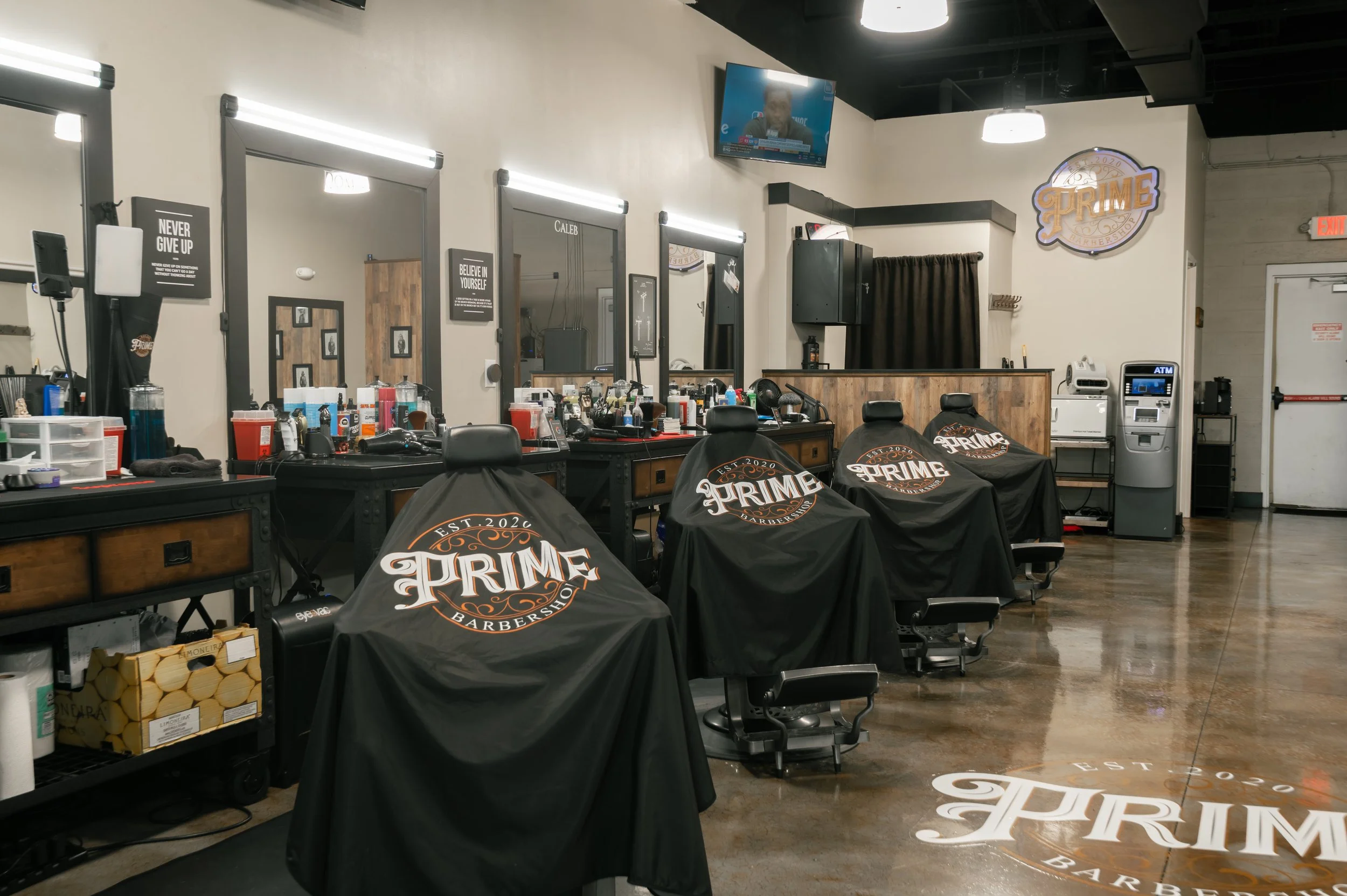 Barber shop with black barber chairs covered in black cloth with the 'Prime Barbershop' logo, workstations with barber tools, mirrors with lights, a TV mounted on the wall, and a reception desk with a black curtain and a sign with the shop's logo.