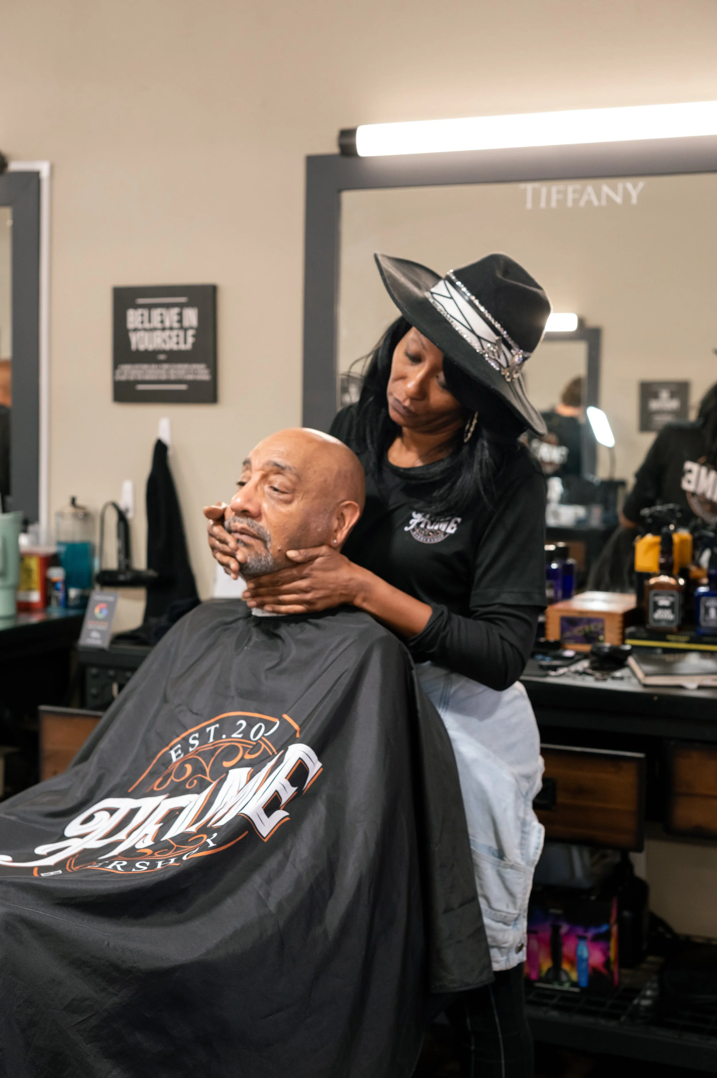 A woman wearing a black hat and earrings is giving a man a shave at a barbershop.