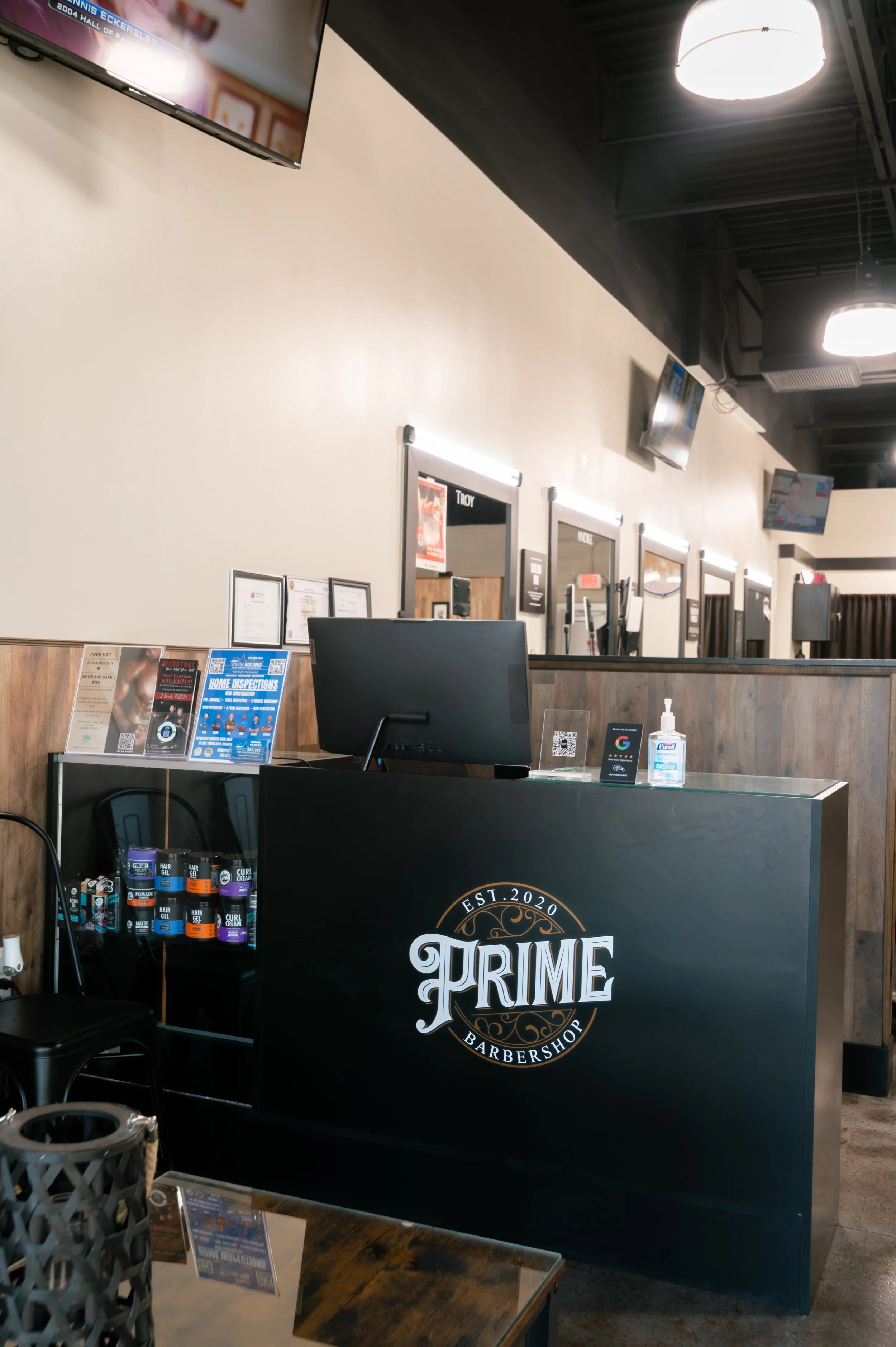 The image shows a reception area of a barbershop called Prime Barbershop, established in 2020. There is a desk with a computer monitor, a bottle of hand sanitizer, and various products such as pomade and hair gel on the counter. Behind the desk, ther