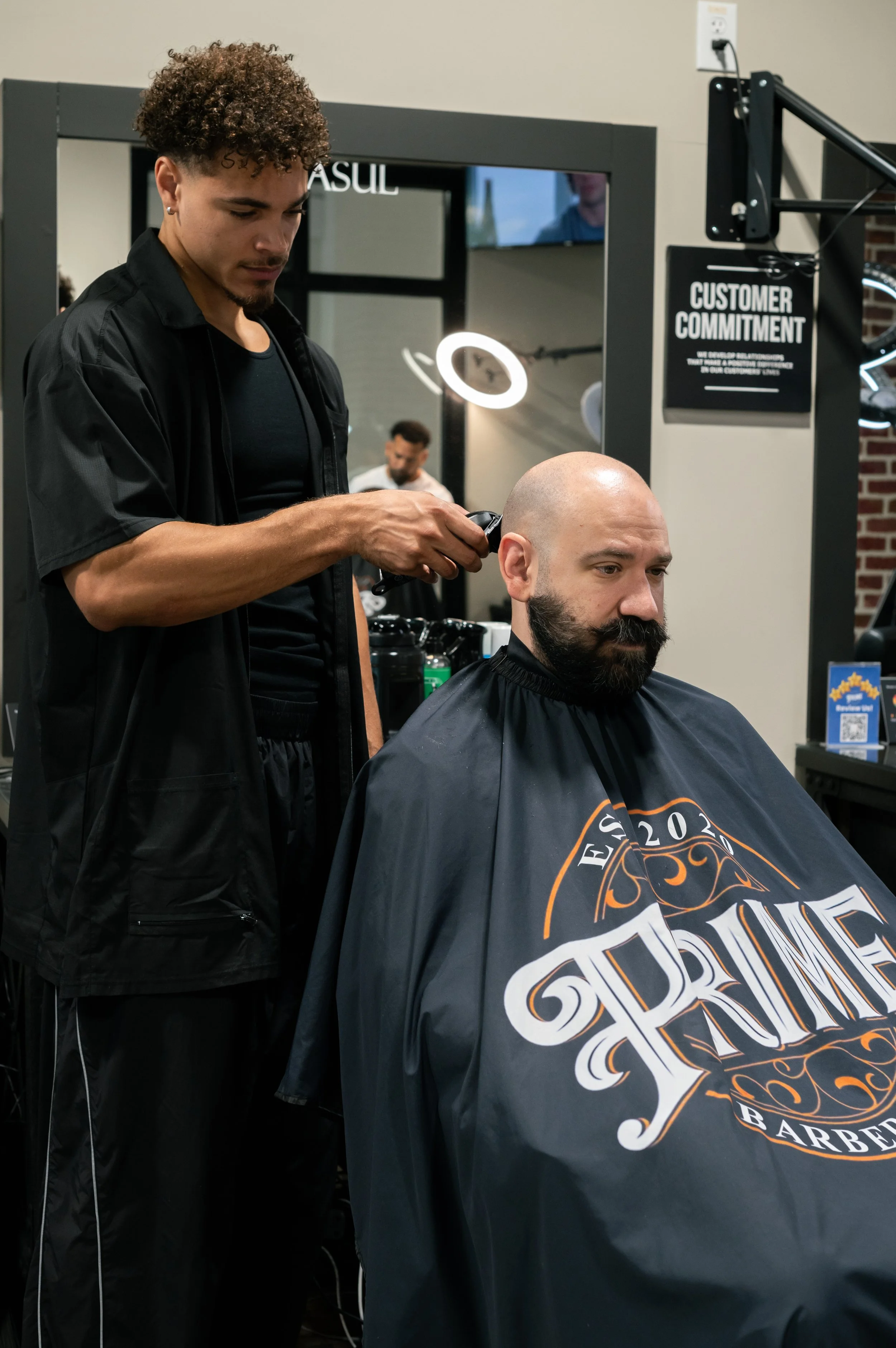 A barber with curly hair cuts a man's beard with clippers in a barbershop. The man has a shaved head and a beard, wearing a barber cape with the words 'Est. 2020' and 'Barber' visible. The background includes a sign that reads 'Customer Commitment' a