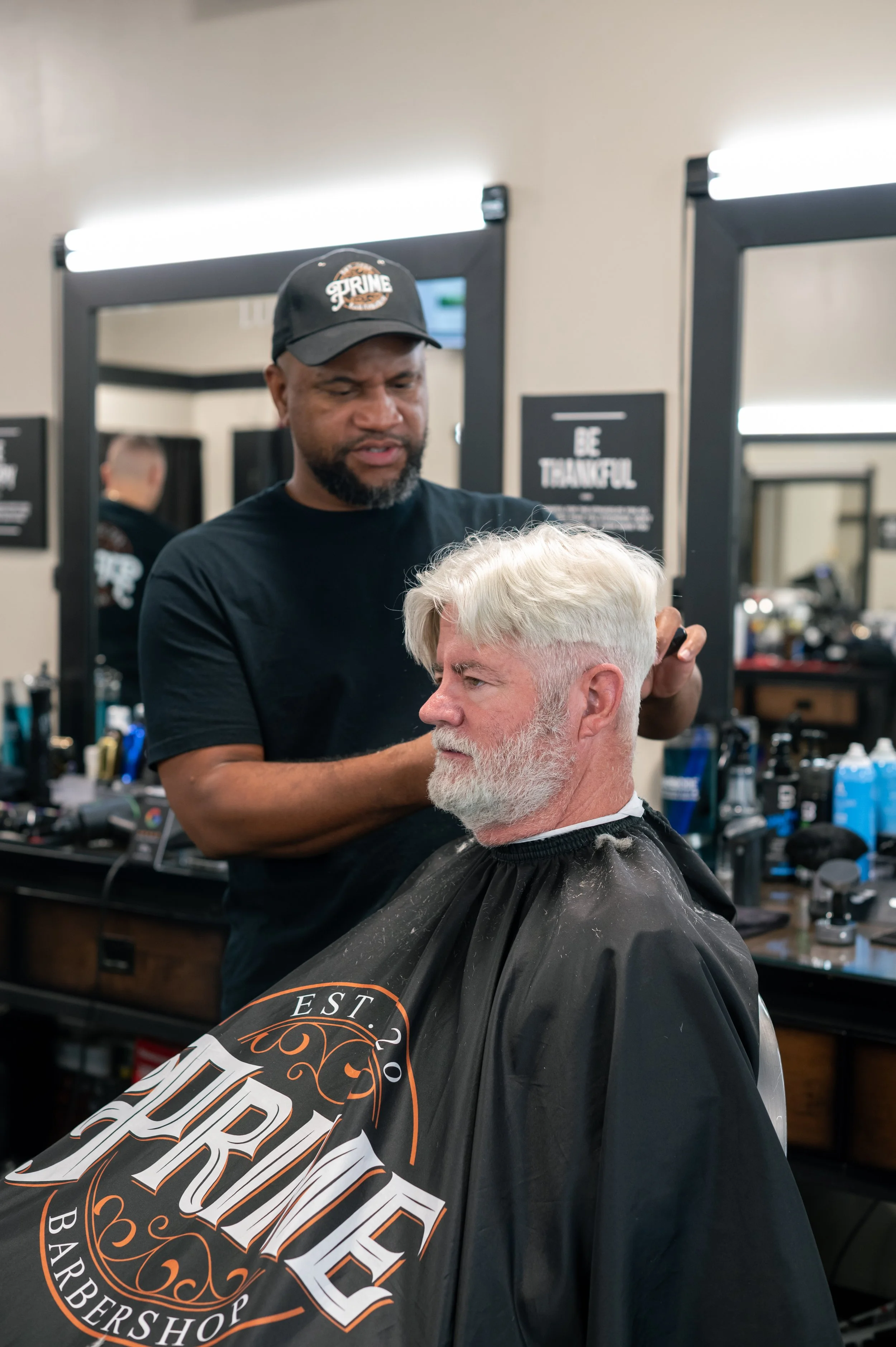 Barber cutting a man's hair in a barbershop with mirrors and tools visible in the background.