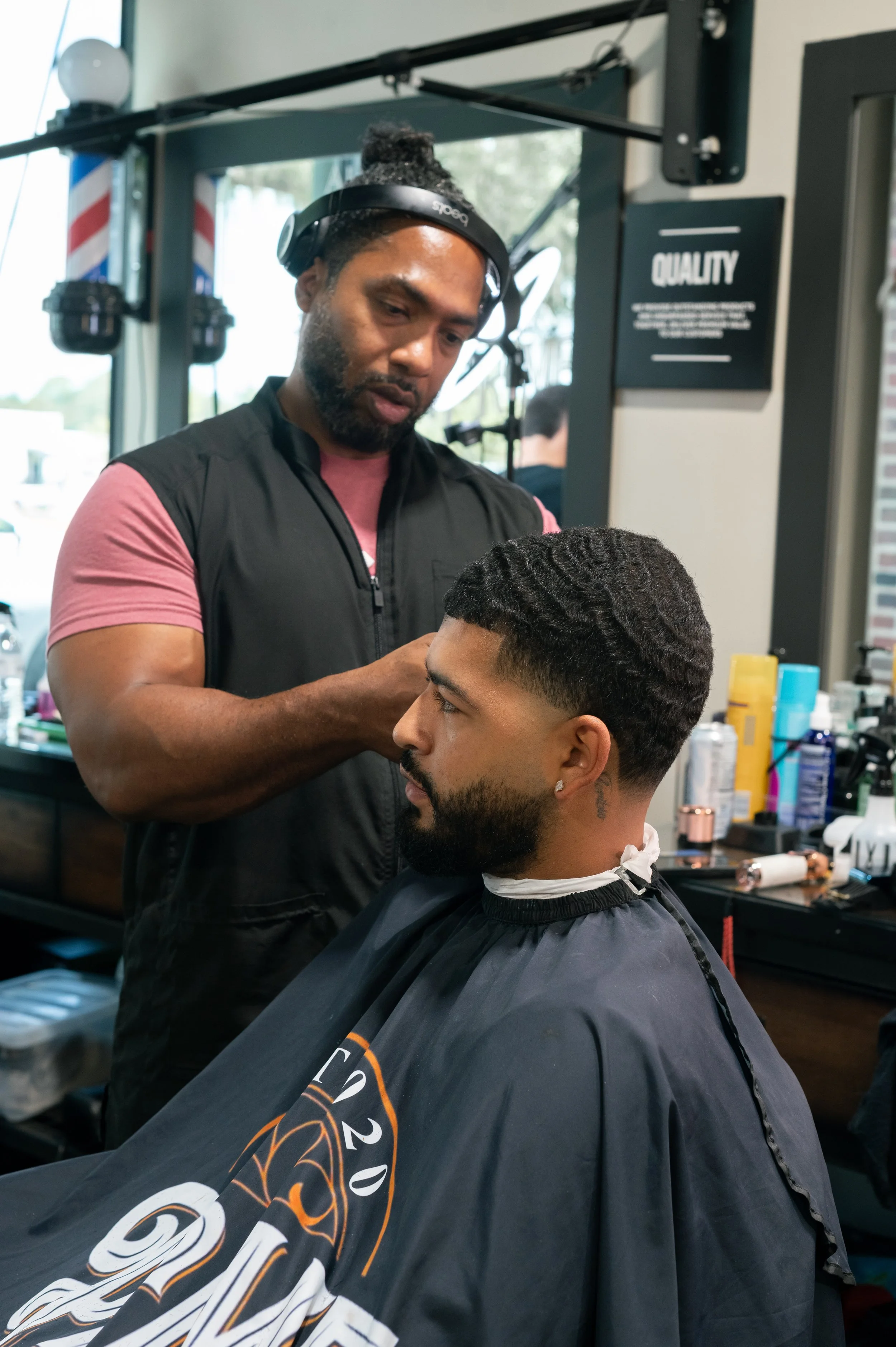 A barber giving a haircut to a male customer with a barber cape in a barbershop, with shelves of grooming products and a sign that reads 'QUALITY' in the background.