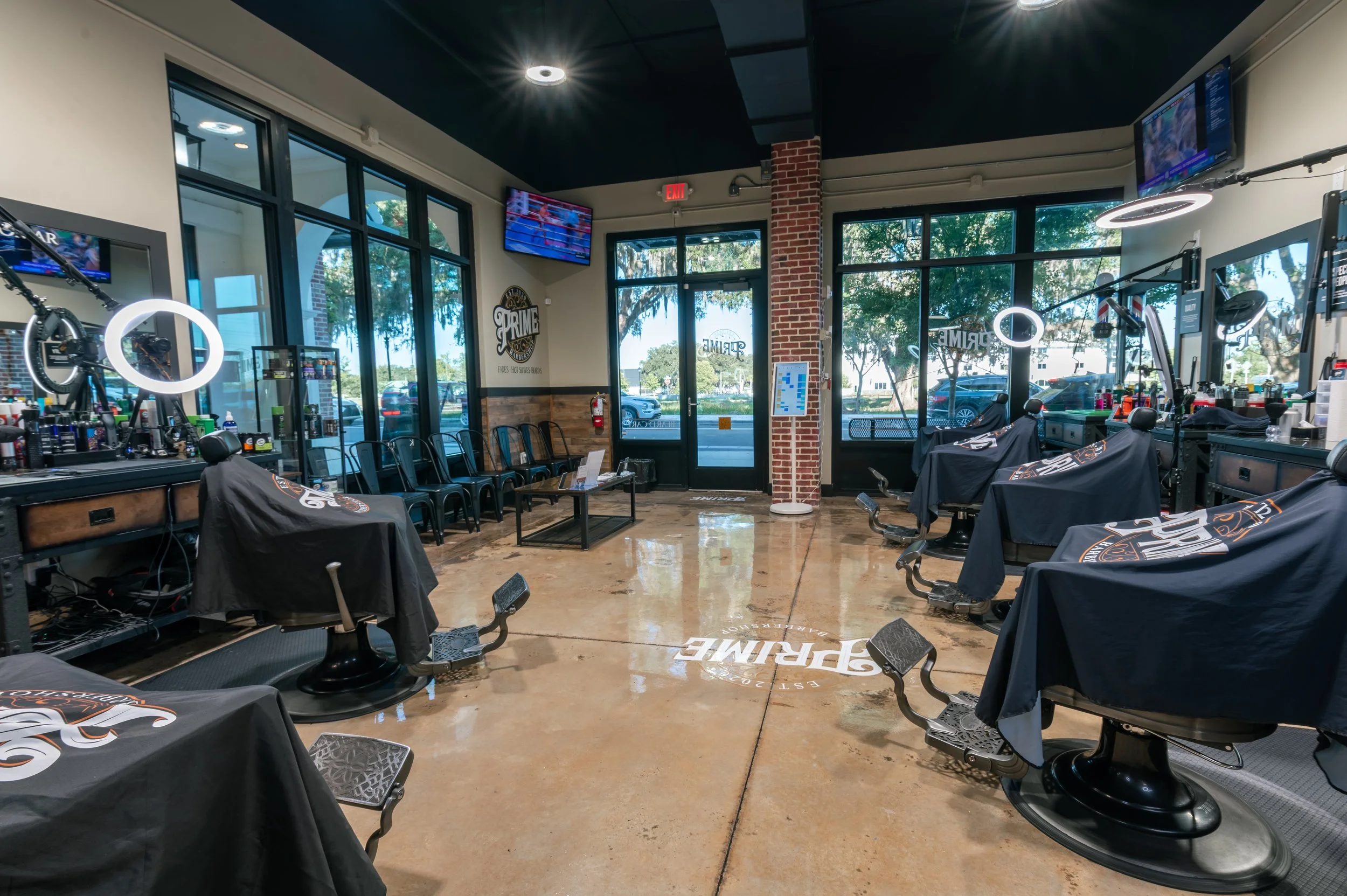 Interior view of a barber or hair salon with black chairs covered in black cloths, large windows letting in natural light, and multiple wall-mounted televisions. There are hair styling stations and tools visible.