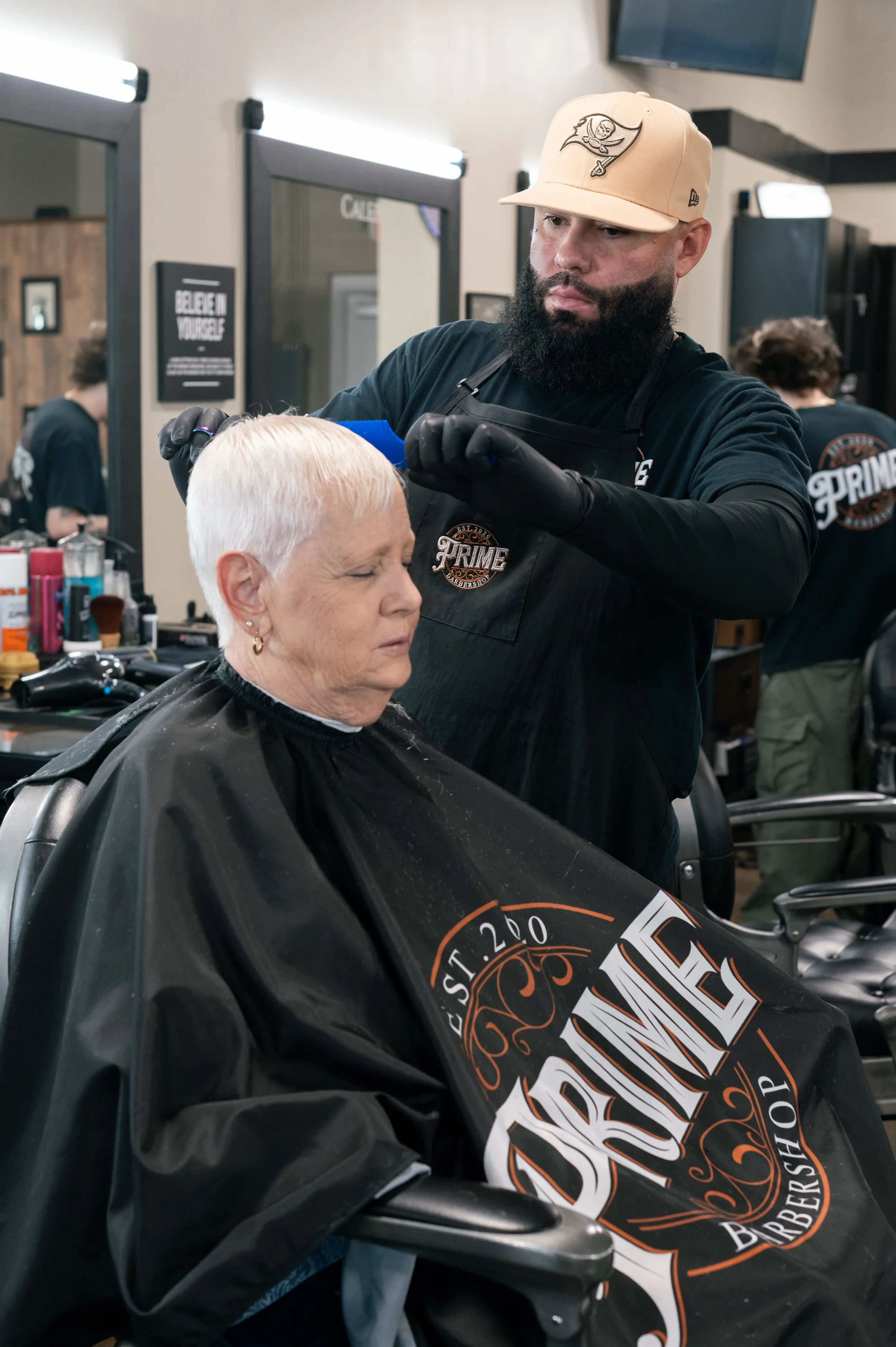 A man getting a haircut at a barbershop, wearing a beige baseball cap, black shirt, and black gloves, while an older woman with short white hair sits in the barber chair with a black cape. There are mirrors and barber supplies in the background.