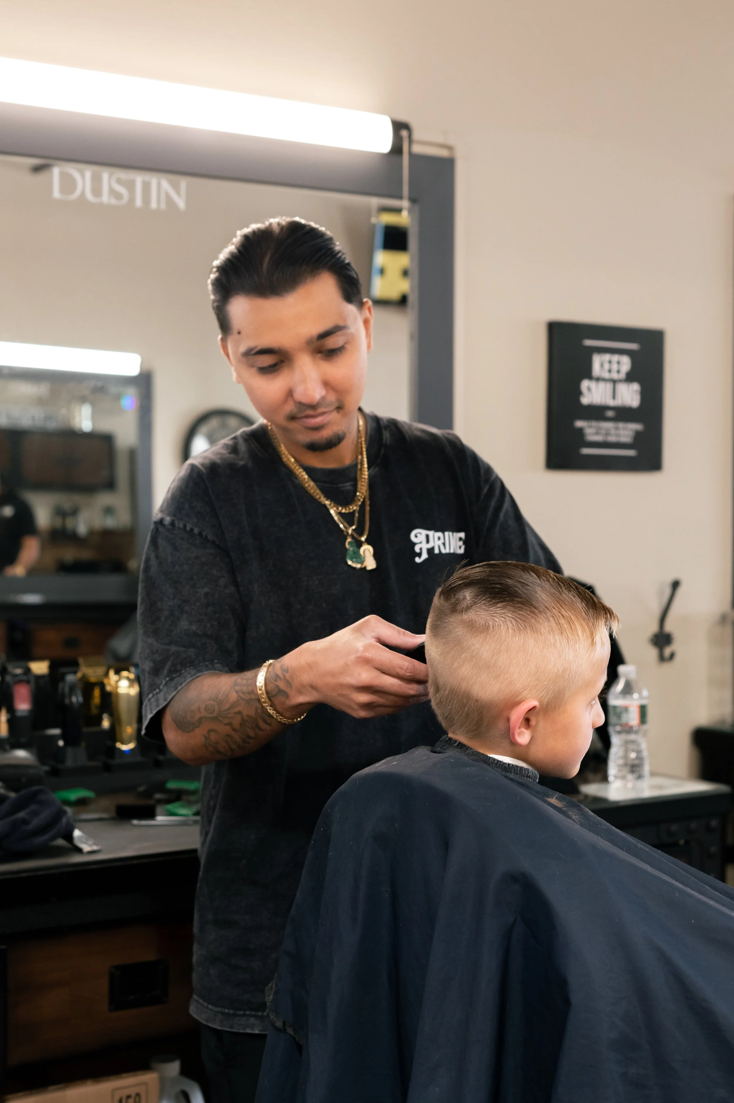A hairstylist with tattoos, wearing a black t-shirt with the word 'PRIME' on it, is cutting a young boy's hair in a barbershop. The boy has blond hair and is wearing a black cape. There is a water bottle, a sign that says 'KEEP SMILING,' and various 