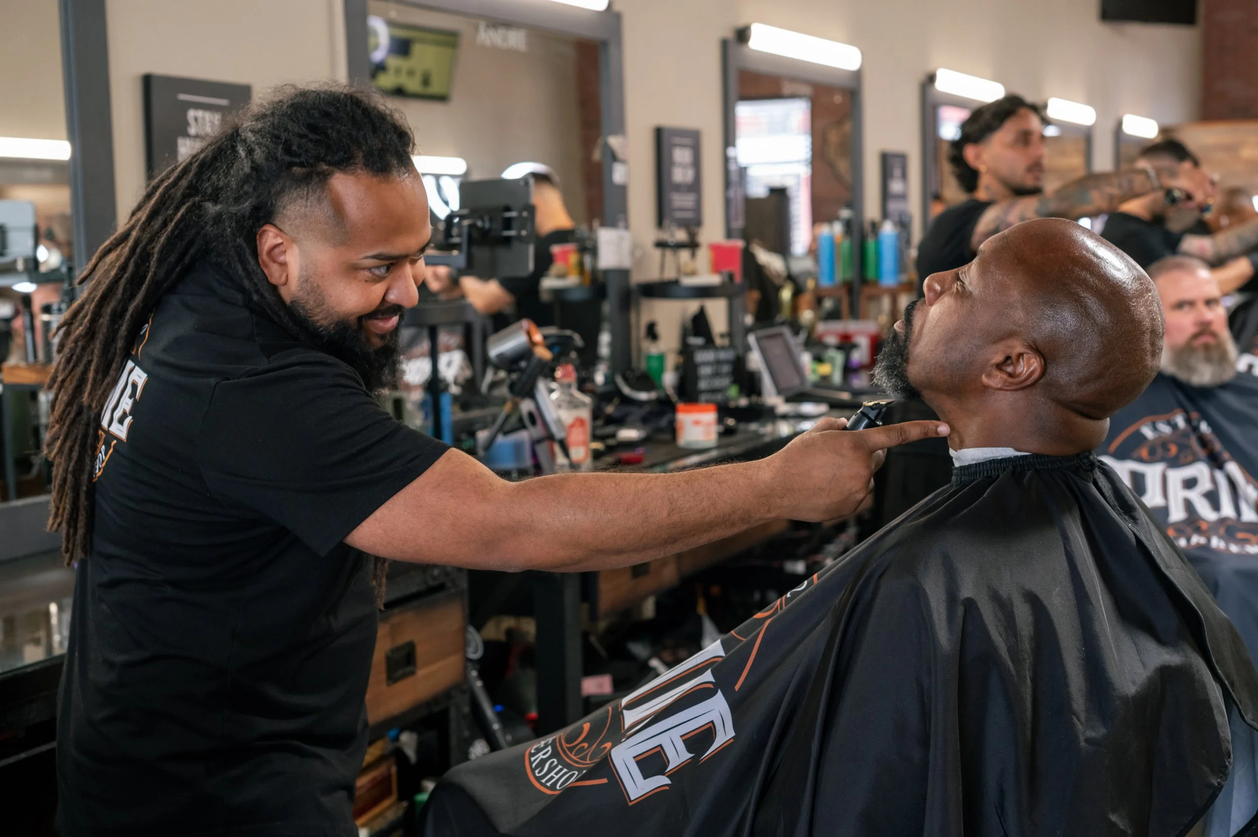 A man with dreadlocks giving a haircut to a customer in a barber shop, with other customers and barbers visible in the background.