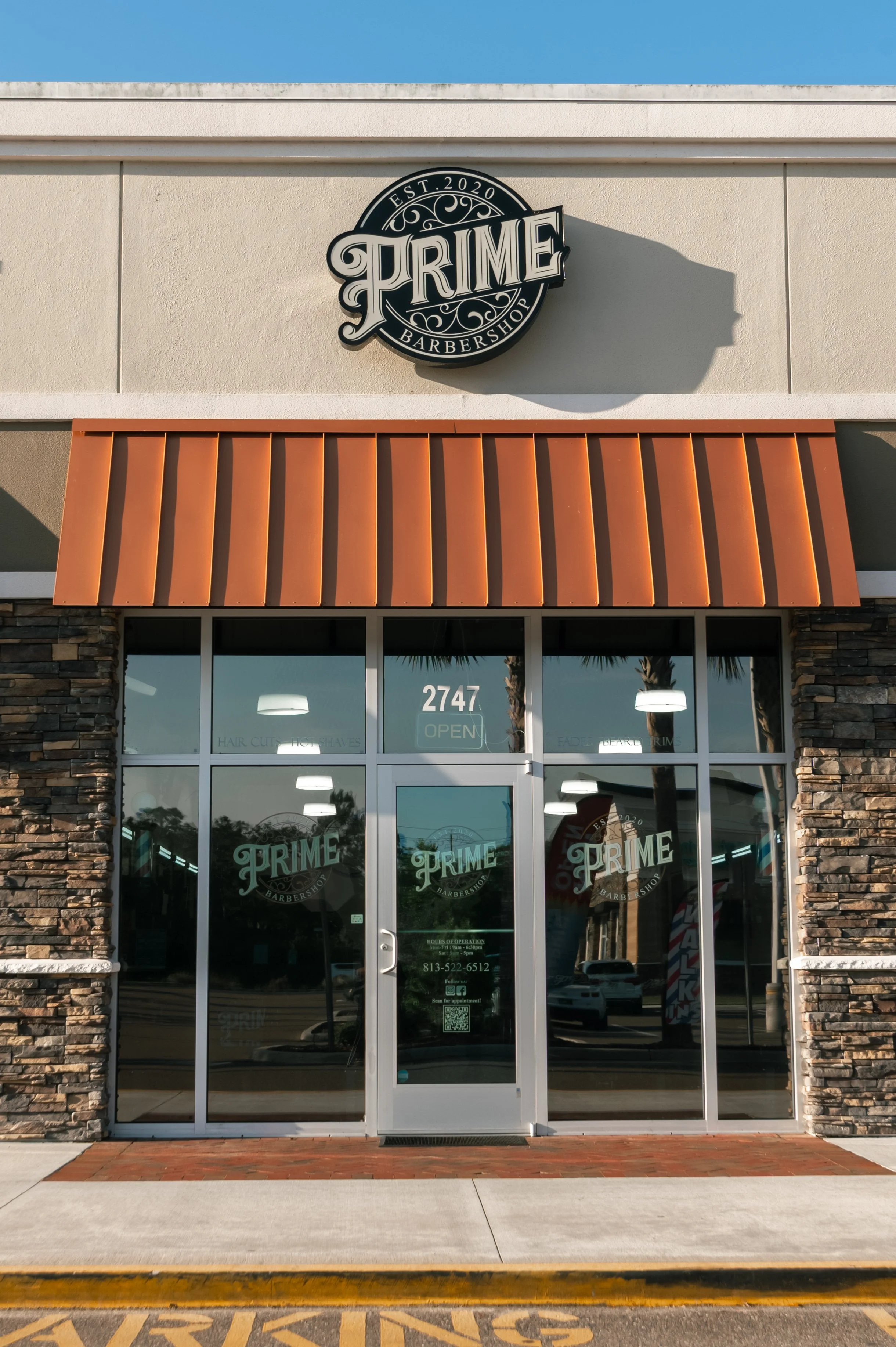 The storefront of Prime Barbershop with a large sign, orange awning, glass doors, and windows at 2747. Palm trees and cars are reflected in the glass. The sign indicates the shop was established in 2020.