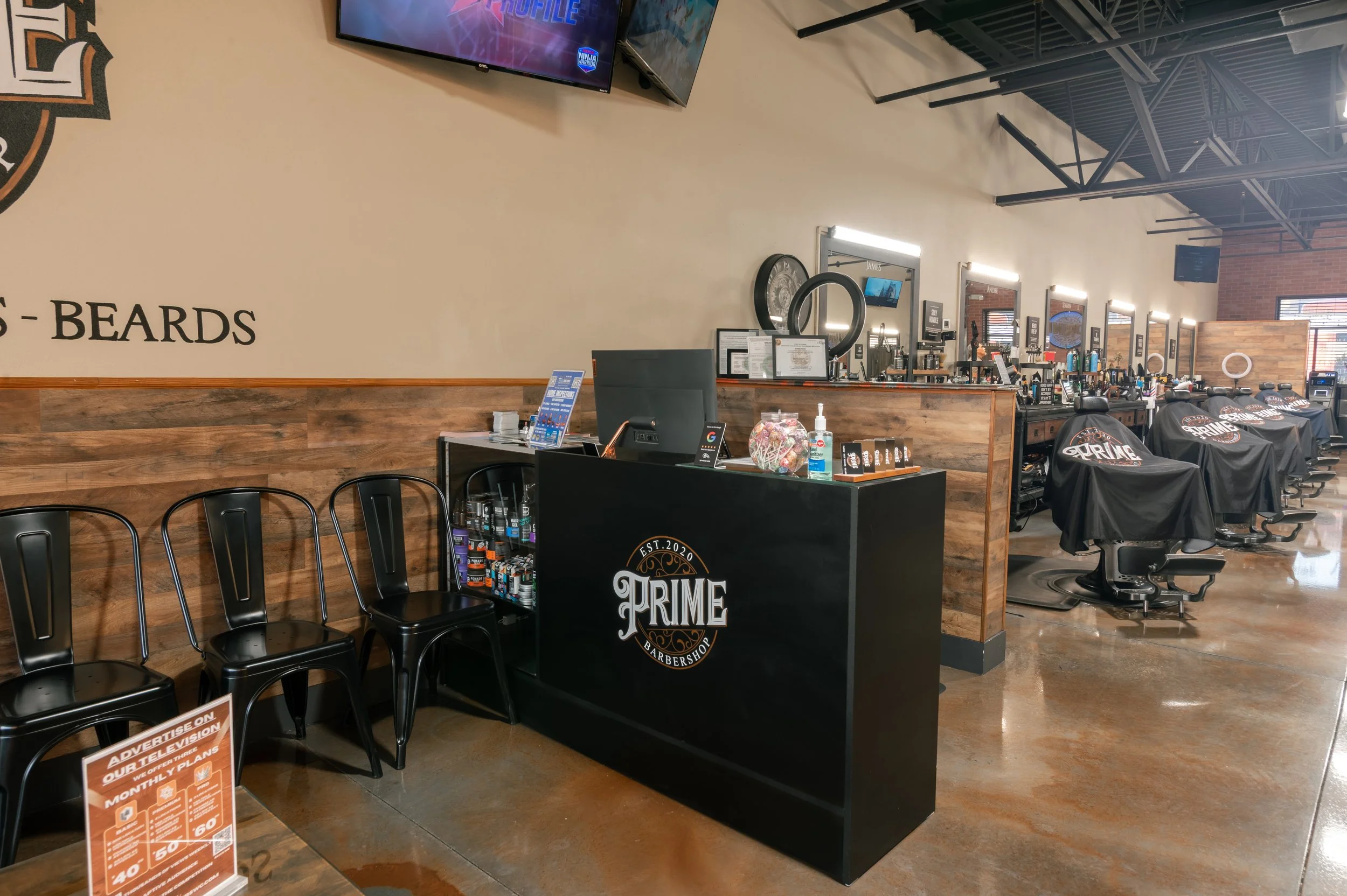 Barbershop interior with black chairs, mirrors with lights, and barber equipment. There is a counter with products, hand sanitizer, and informational signage. The wall features wood paneling and signage indicating 'BEARDS' and the barbershop's logo '