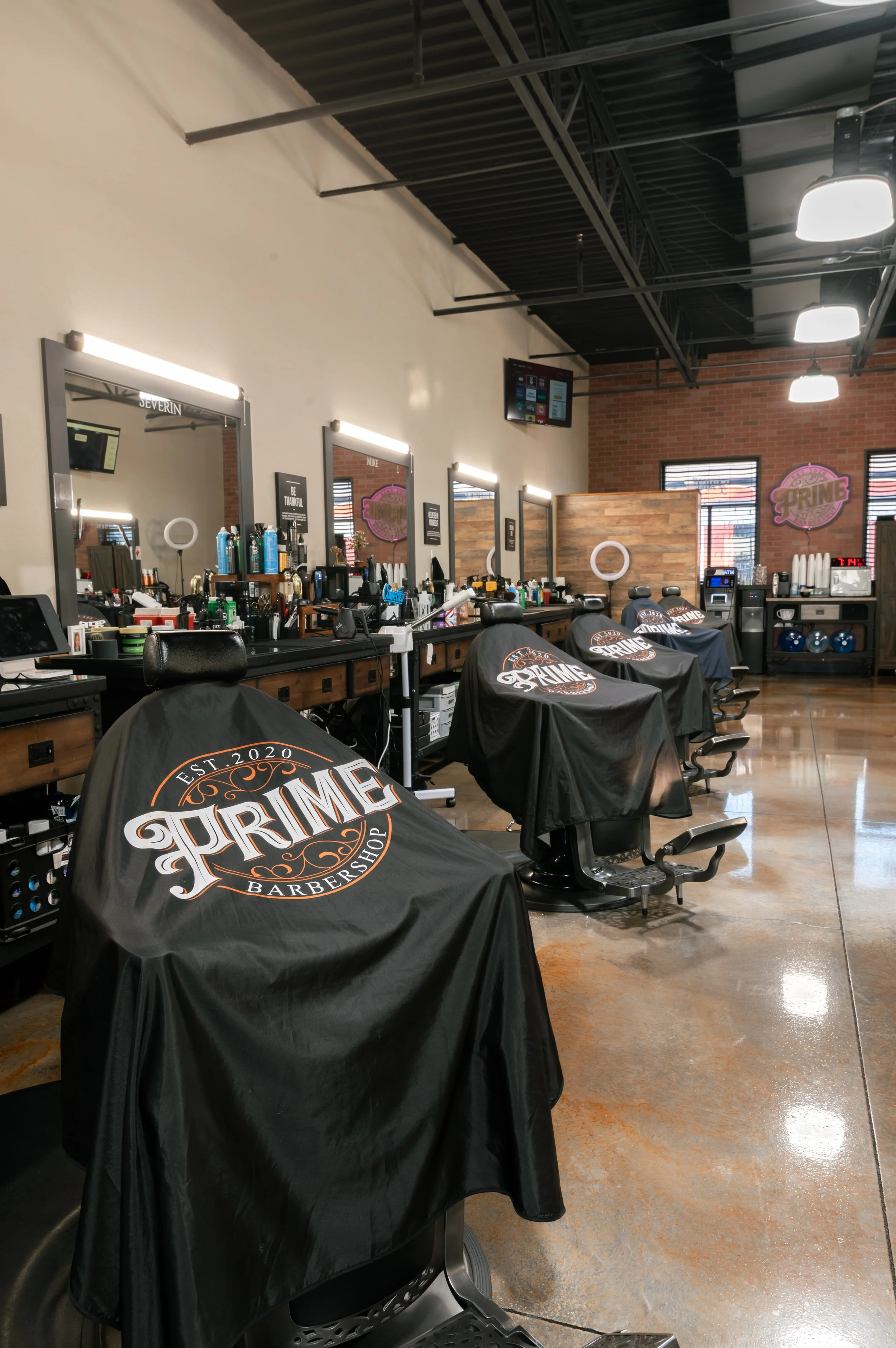 Interior of a barbershop with several styling stations, each equipped with mirrors, barber chairs covered with black capes displaying the 'Prime Barbershop' logo, and various barber tools and products on the countertops. The space features industrial