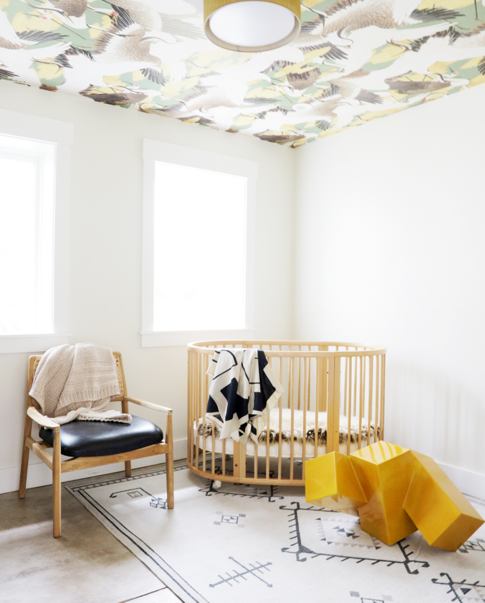 Nursery with crane and cloud motif wallpaper, a gold flush-mount light, round wood spindle crib, cane-and-leather accent chair, and patterned rug, Oakland Bungalow (61st Street) end-to-end remodel by interior design studio Spearman Spaces