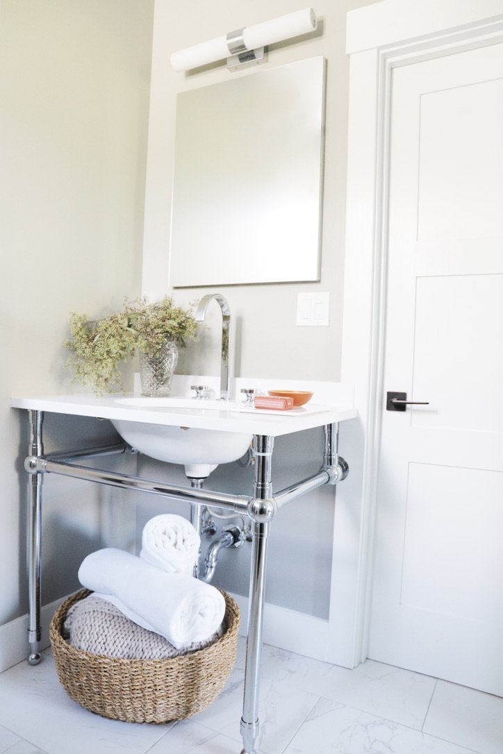 White bathroom vanity with a chrome console frame, marble top, gooseneck faucet, a crystal vase of dried flowers, and a woven basket of rolled white towels beneath, Oakland Bungalow (61st Street) full-service remodel by Spearman Spaces