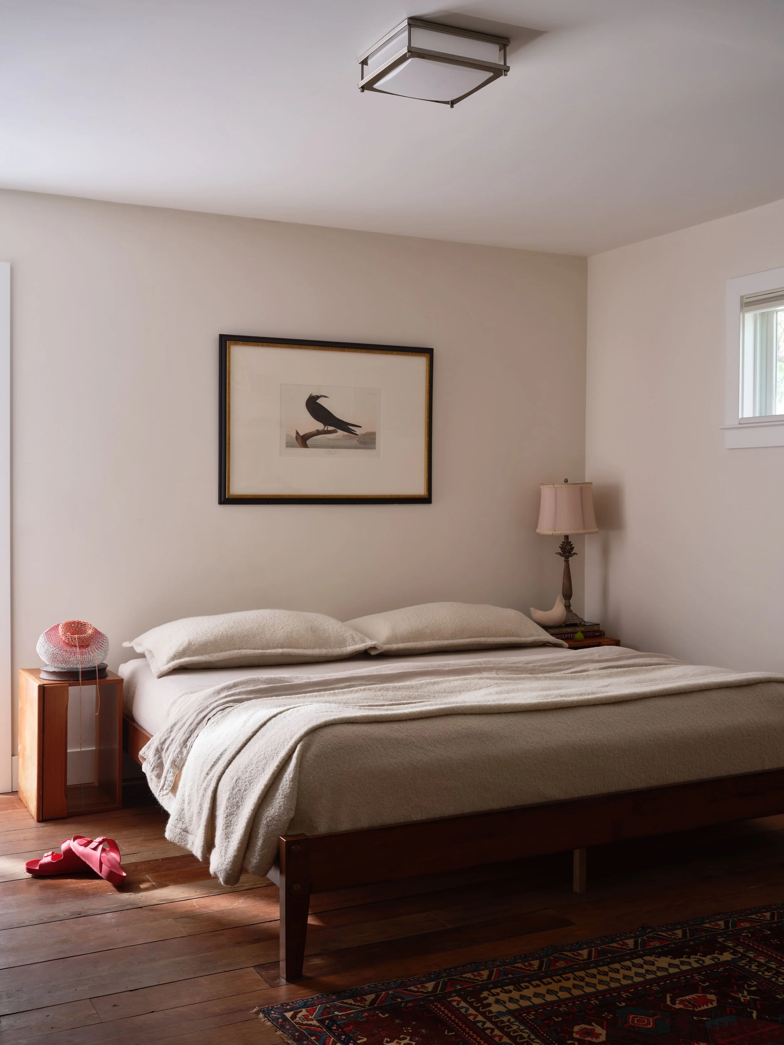Bedroom with a framed bird print above a wood platform bed dressed in oatmeal bedding, a square flush-mount ceiling light, a wood nightstand with a woven fringed table lamp, a patterned vintage rug at the foot of the bed, and red slide sandals on wid