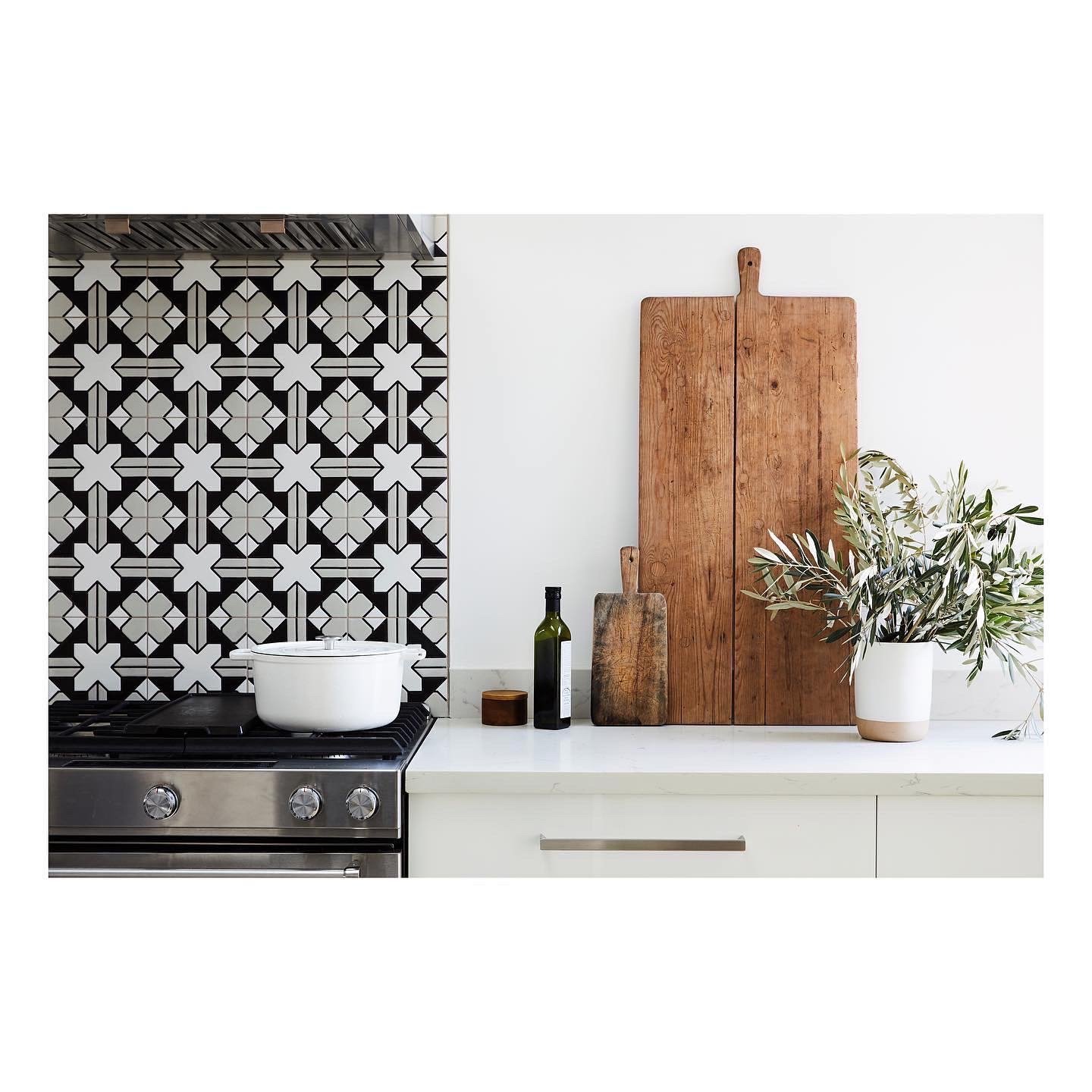 Kitchen detail with a black and white graphic cross-pattern tile backsplash, a black range with a white enameled Dutch oven, layered vintage wood cutting boards, and an olive branch in a ceramic vase on a white marble counter, San Francisco Craftsman